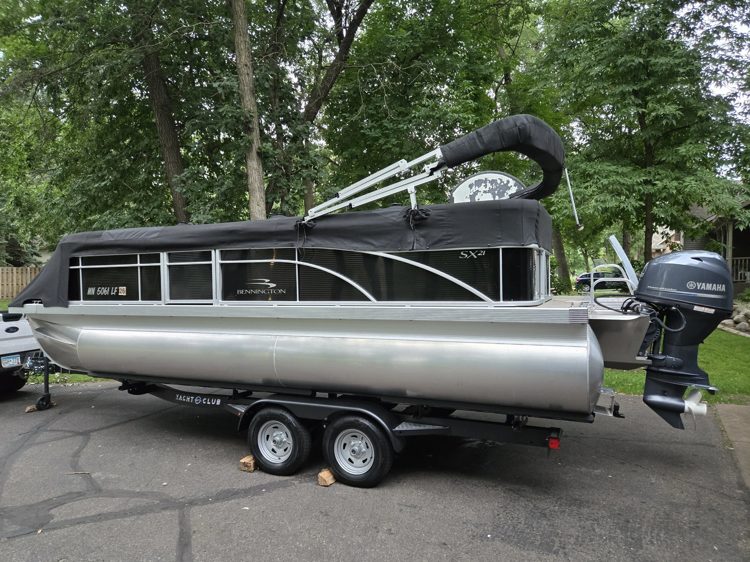 A pontoon boat on a trailer parked on a driveway. The boat has a black canopy and a Yamaha outboard motor. Surrounding trees and a house are visible in the background.