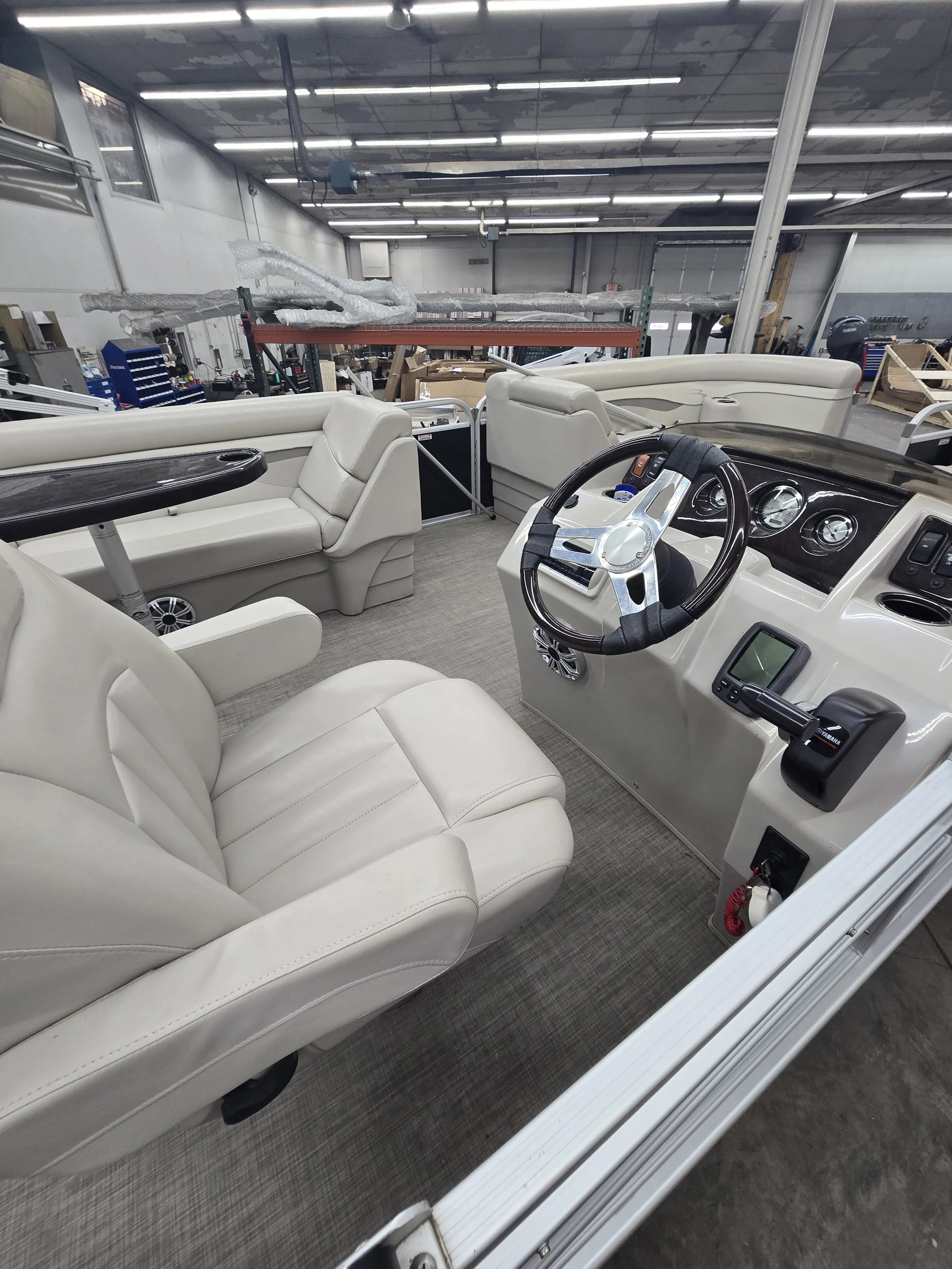 Interior of a pontoon boat with white leather seats, dashboard with gauges, and a steering wheel, inside a warehouse with tools and equipment. Depth finder and Bluetooth radio included