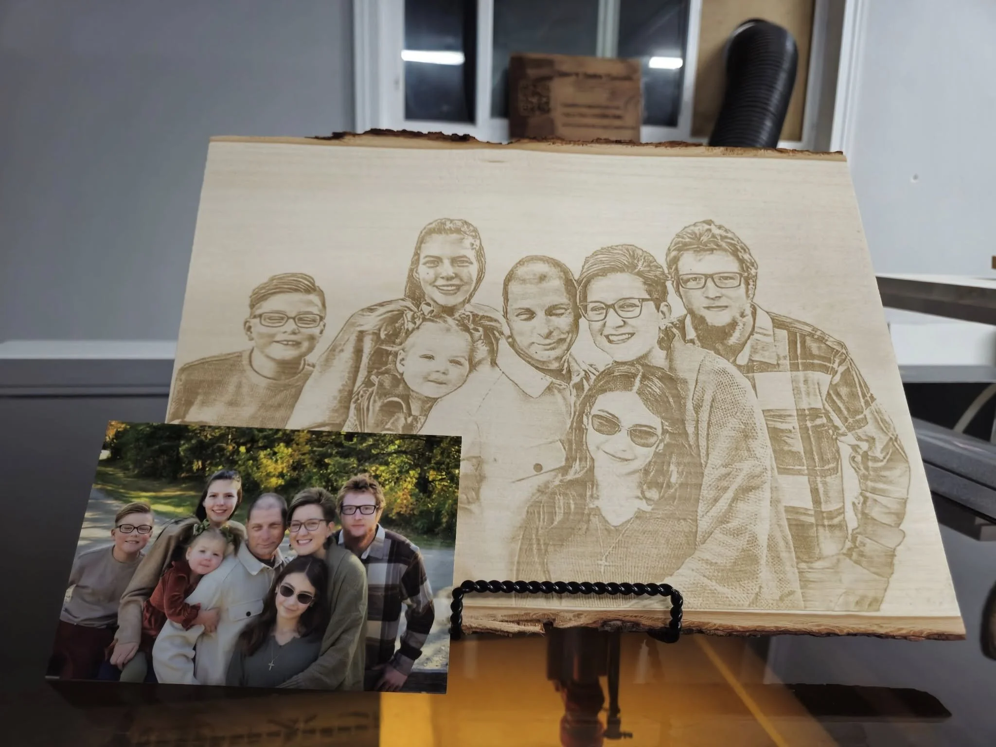 A photo of a family is placed in front of a wooden plaque. The photo shows several people smiling and posing outdoors. The wooden plaque behind it has a laser-etched replica of the same family image. The background features windows and a workshop set
