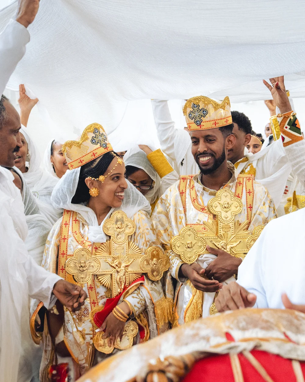 Couple in traditional attire holding golden crosses, surrounded by guests in white