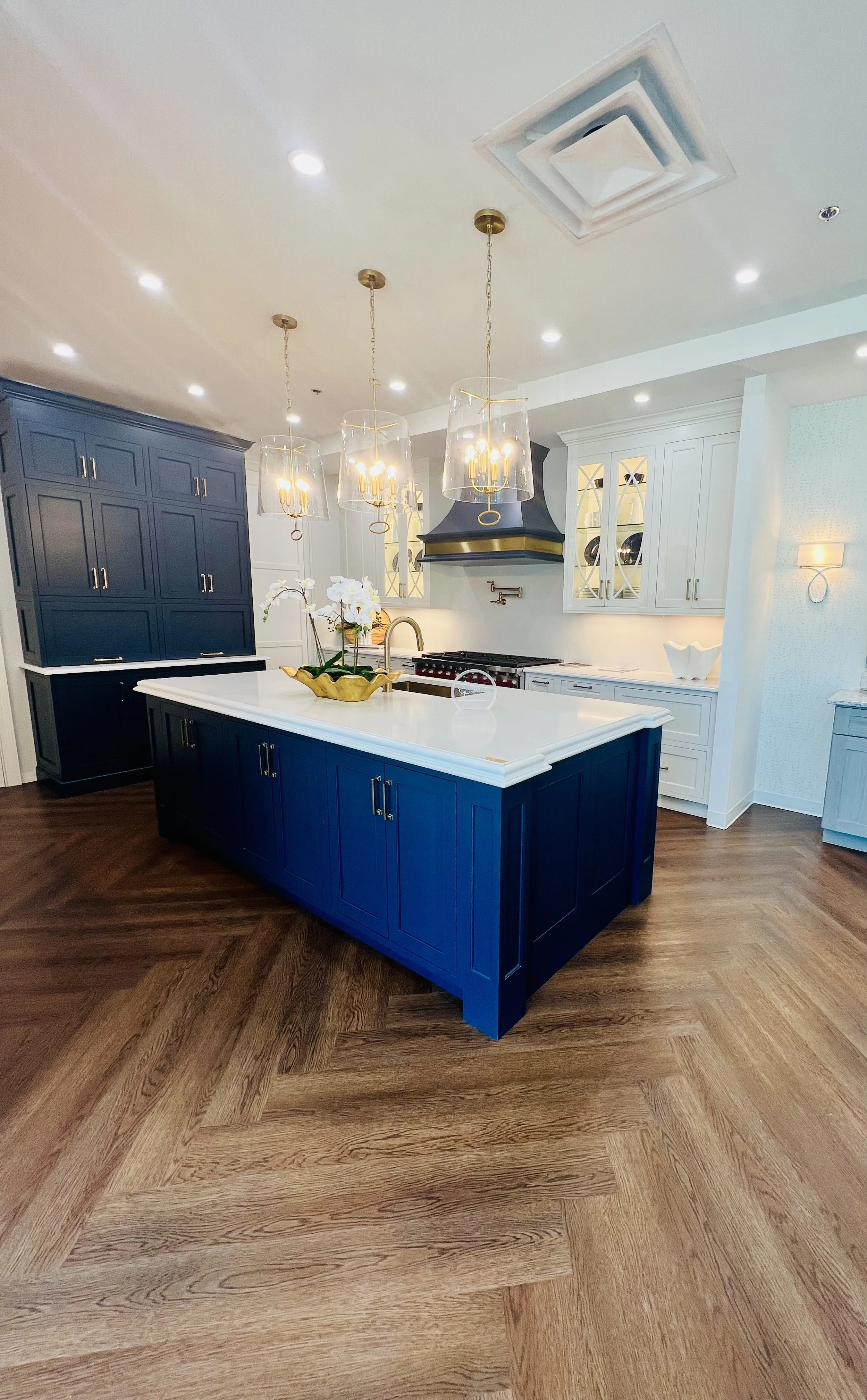 A modern kitchen with a navy blue island and matching tall cabinets, white countertops, white upper cabinets, and gold accents. There are three pendant lights above the island, and the kitchen features hardwood flooring.