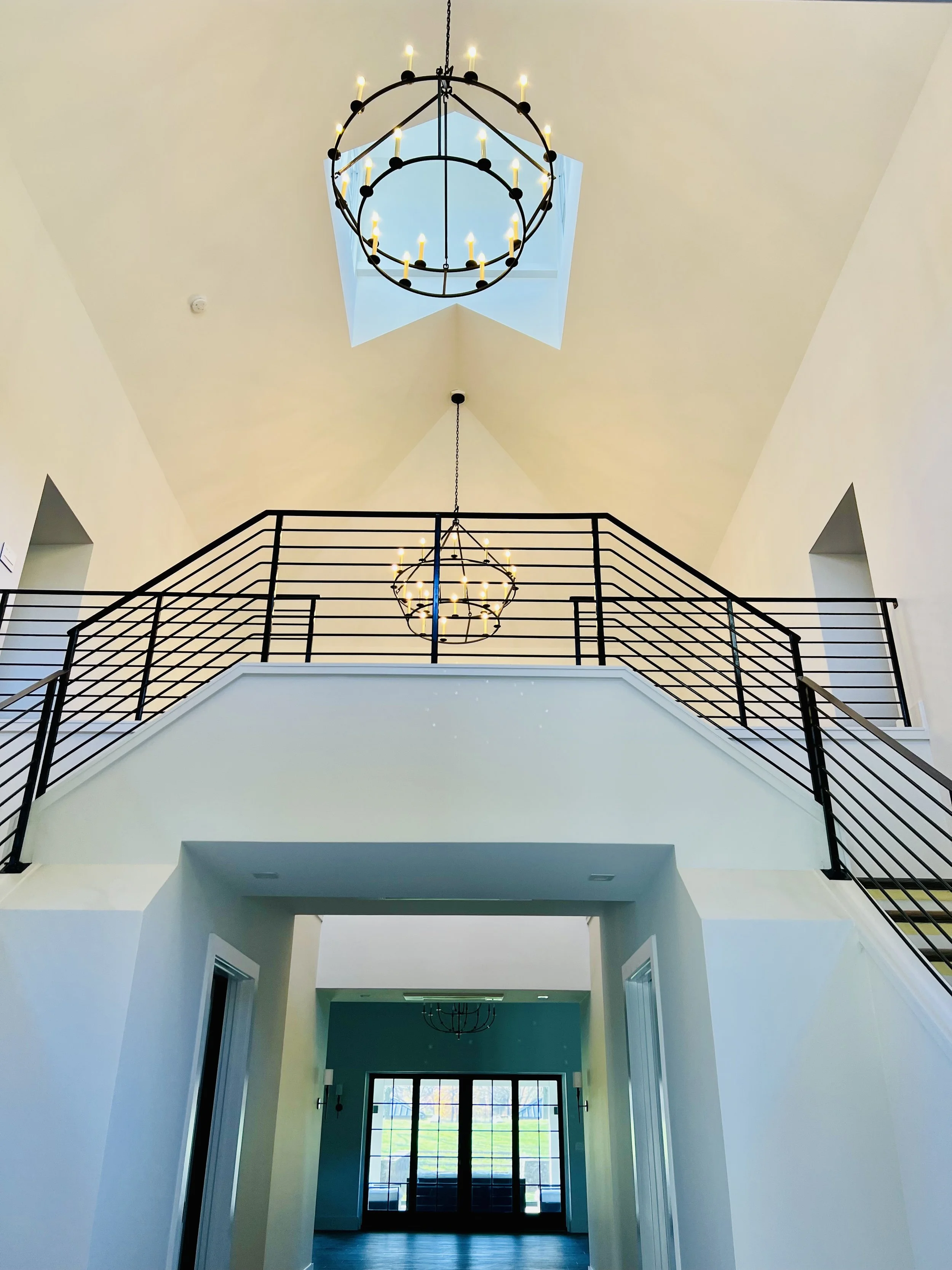 Interior view of a modern house foyer with a high ceiling, large windows, and chandelier lighting fixtures.