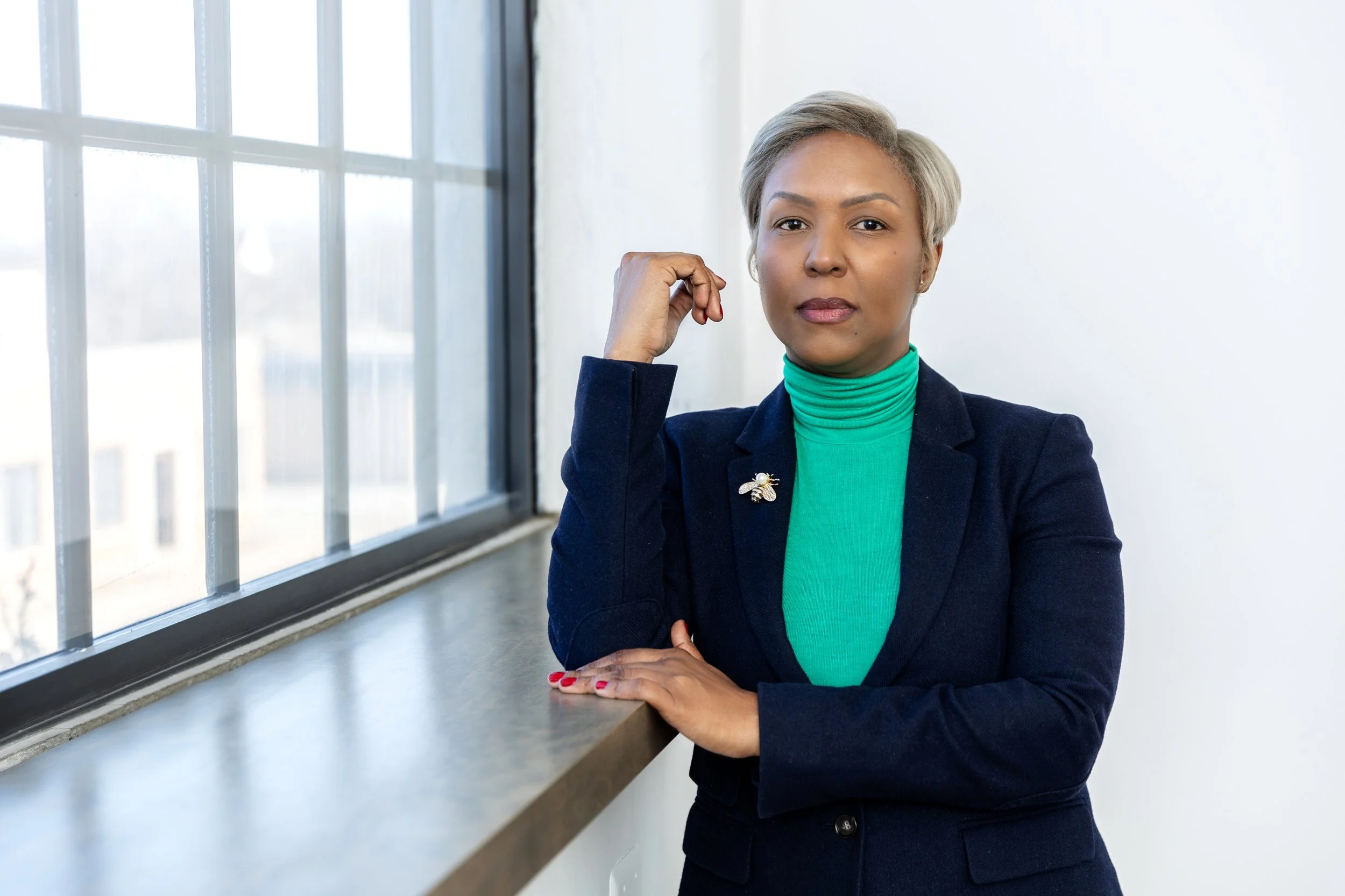 Melinda B. Powers, small business consultant in Detroit, MI. Image is of beautiful black business woman wearing a bright green turtleneck, navy blazer and brooch on her lapel. She is next to a window. Photo by LeoSage Images in Detroit, MI.