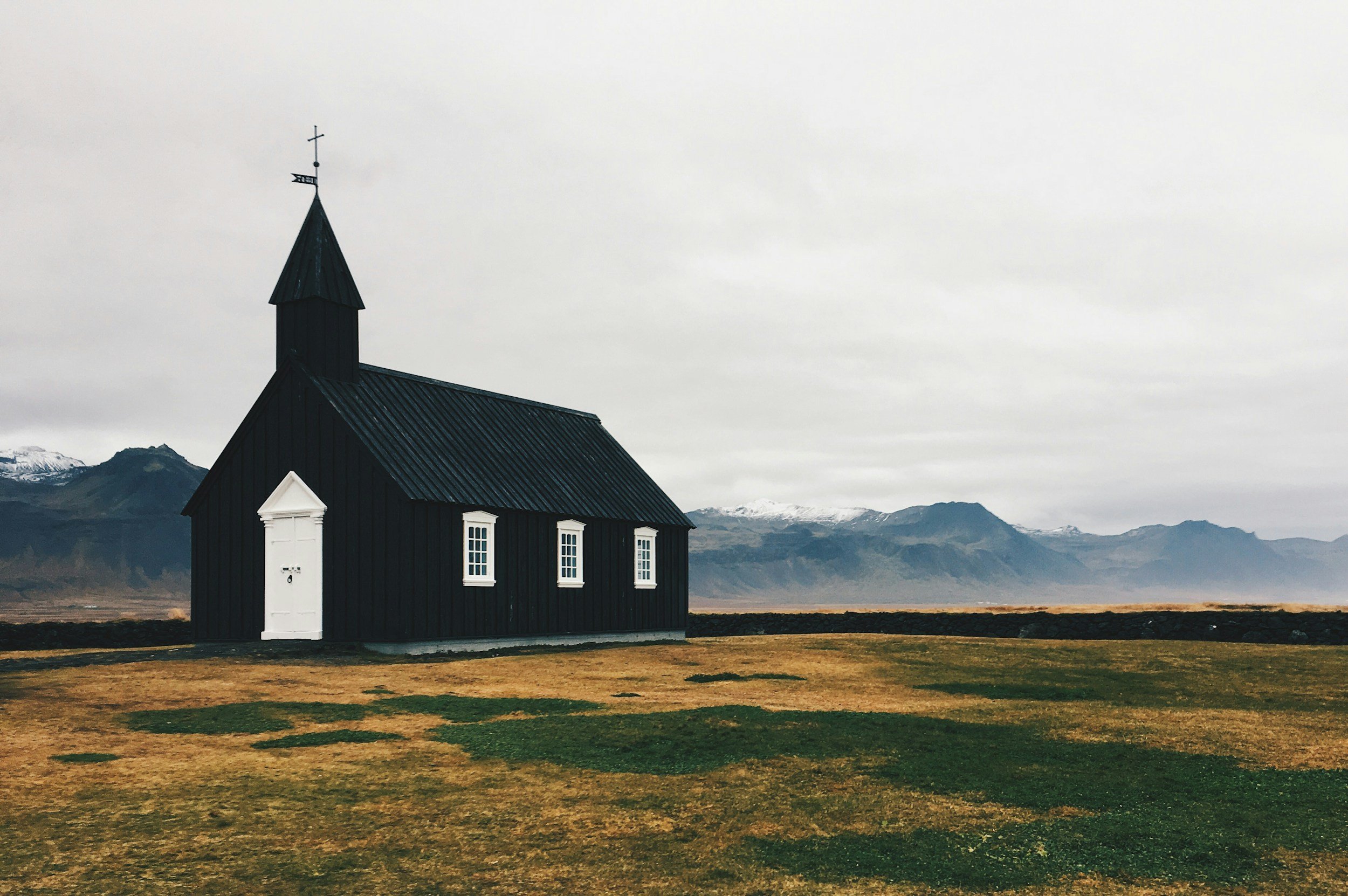 A black church with white windows and door, set in a vast landscape with mountains and snow, overcast sky.
