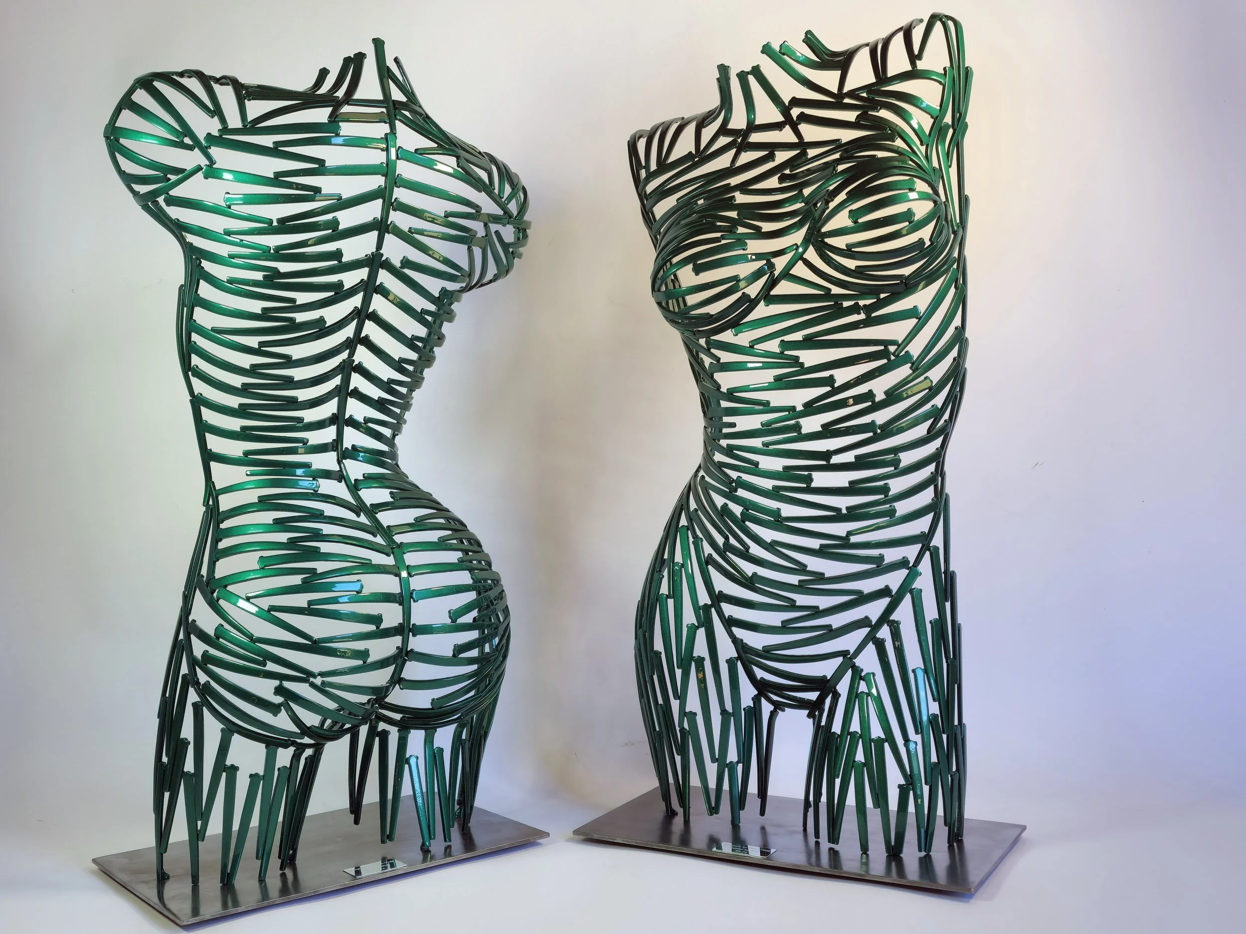 Two metal sculptures of female torsos made from green neon light tubes arranged to outline the shape of the body, with one representing a figure with more pronounced curves and the other a slightly slimmer form.