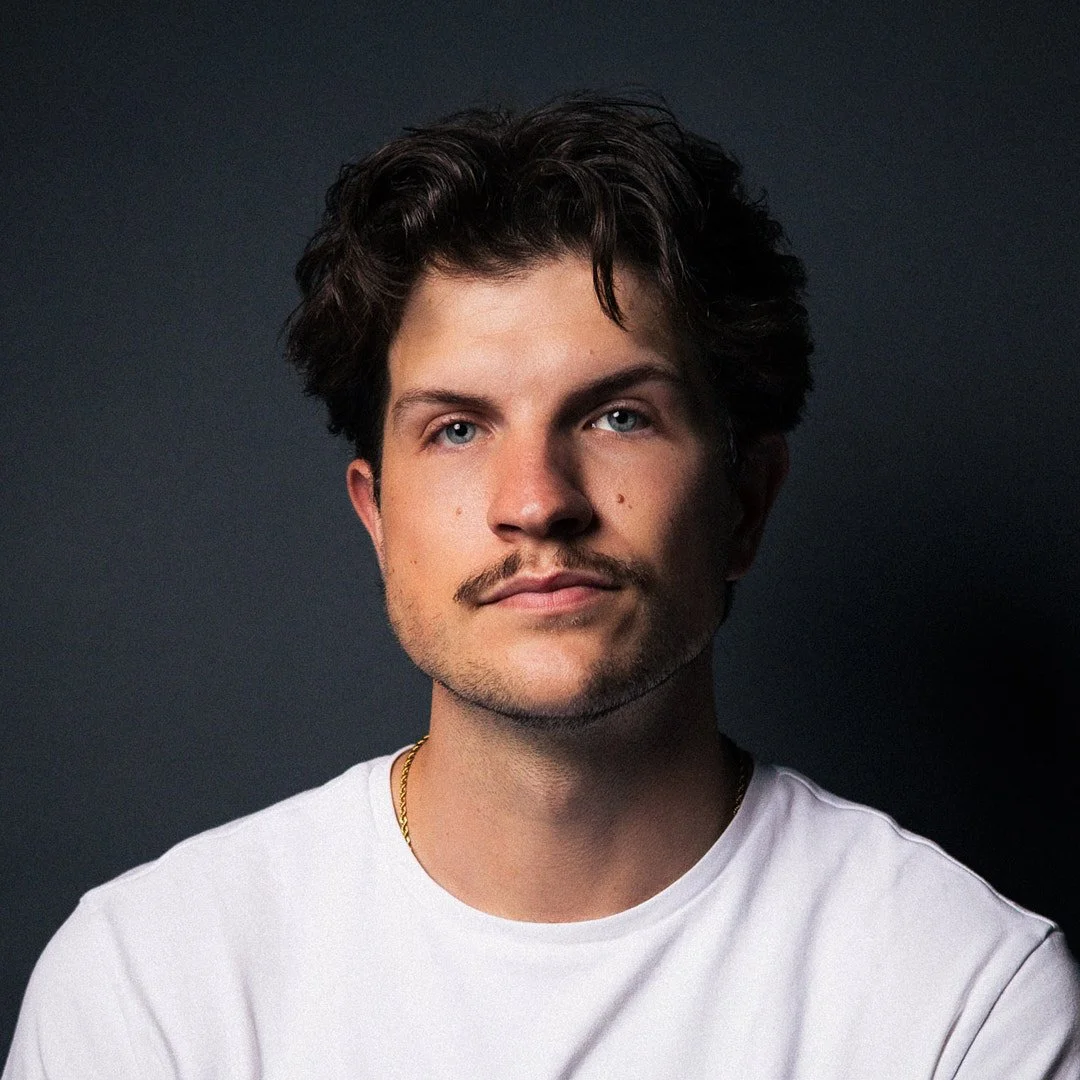 Portrait of a young man with dark hair, blue eyes, and a slight mustache, wearing a white t-shirt against a dark background.