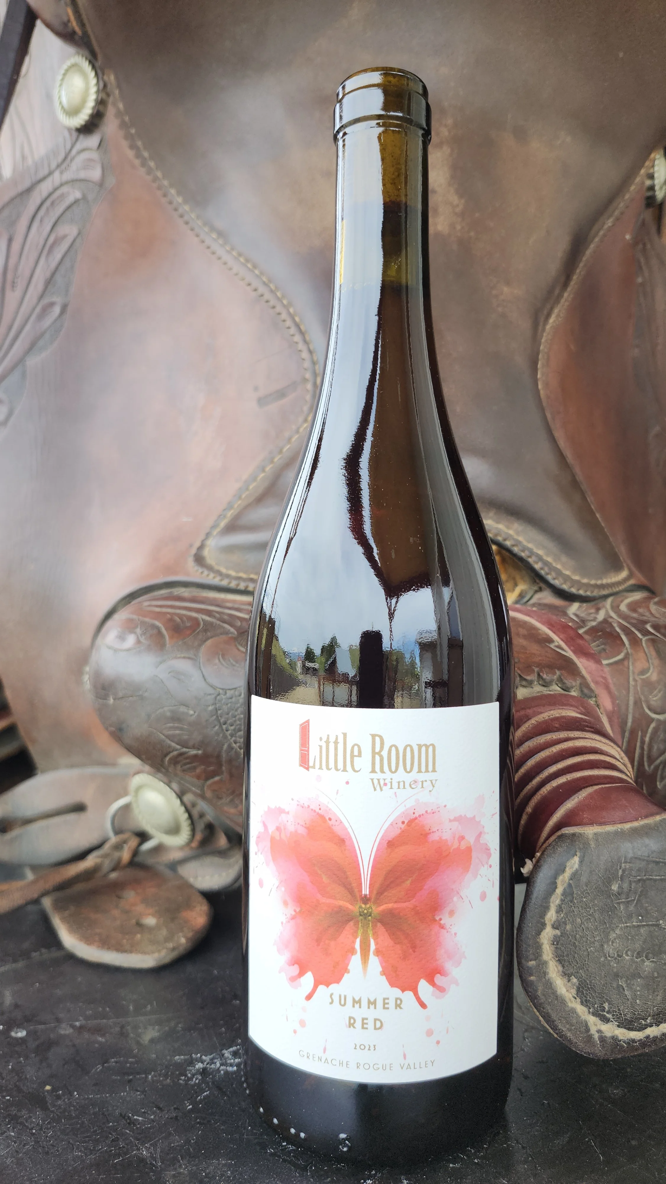 A bottle of Little Room Winery Summer Red wine made from Grenache grapes. Rogue Valley vintage 2023, with a label featuring a pink and orange butterfly, placed on a rustic wooden surface next to a leather horse saddle.