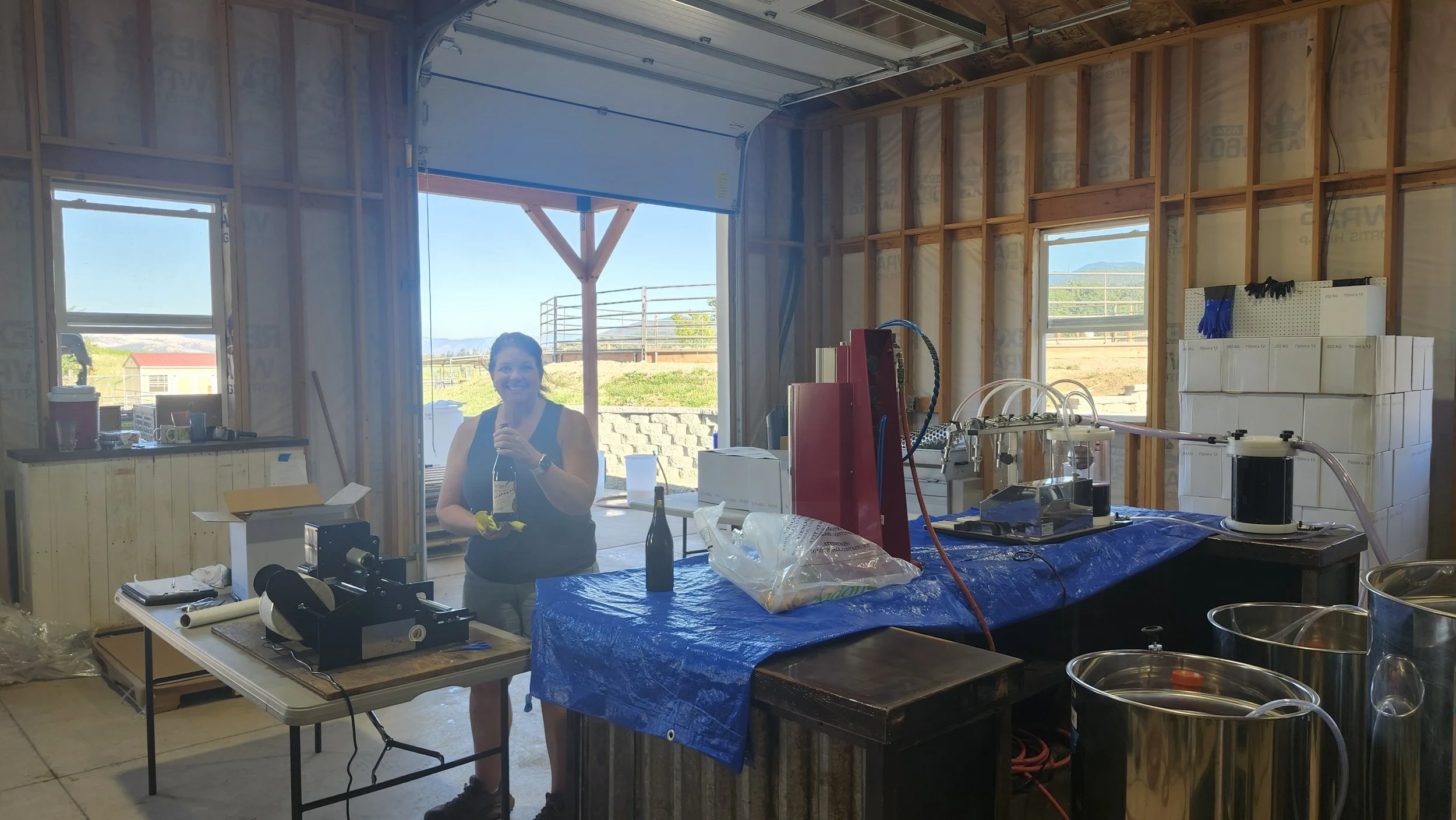 A woman with short dark hair smiling, holding a wine bottle, inside a winery with exposed wood framing and large windows, with outdoor scenery visible.