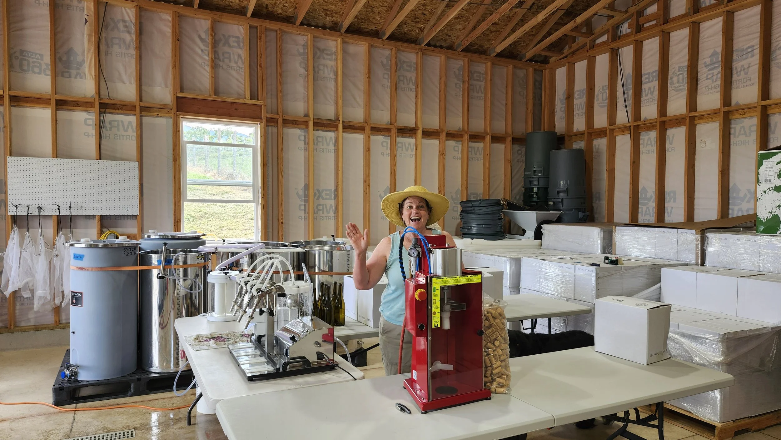 A woman in a yellow hat smiling and waving inside a winery building with exposed wooden framing, work tables, and equipment.