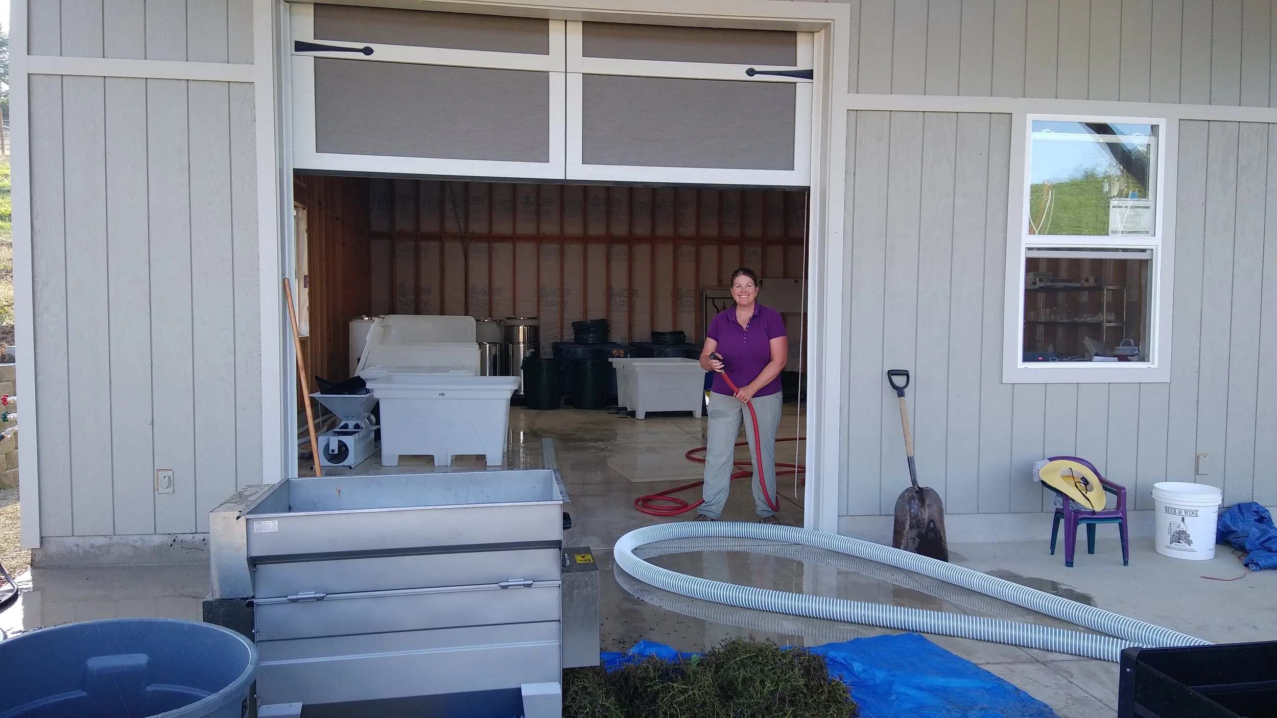 A woman in a purple shirt and beige pants standing inside a winery, she is smiling and is surrounded by wine production equipment