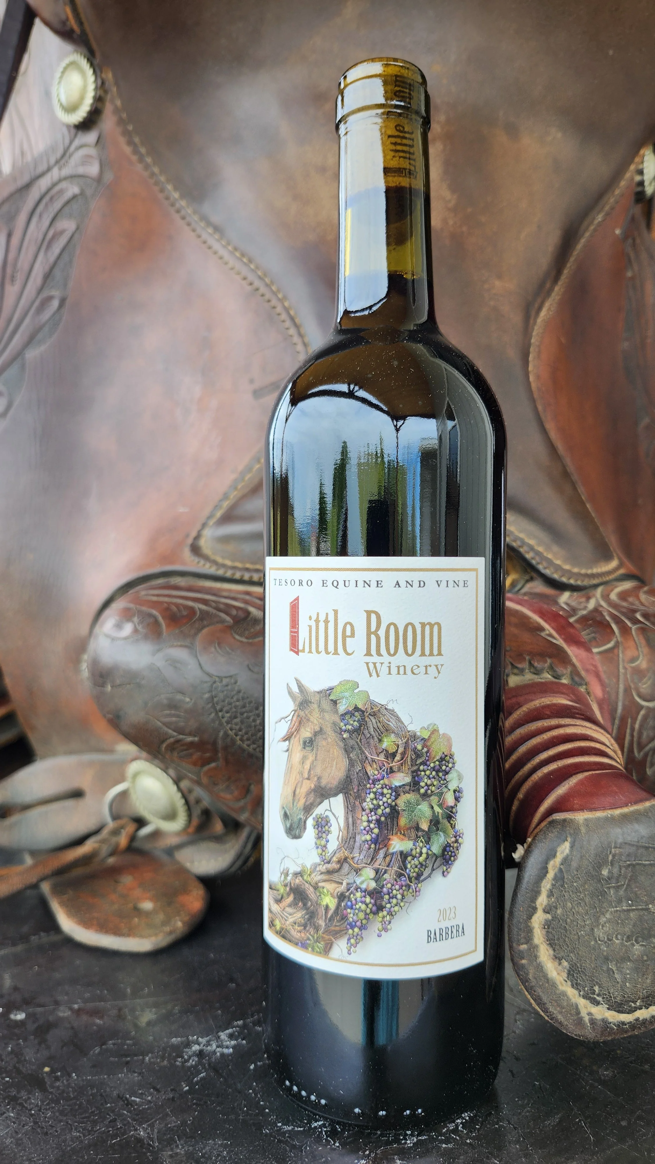 A wine bottle with a label featuring a horse's head and grapevine artwork, placed on a wooden surface with a leather saddle in the background.