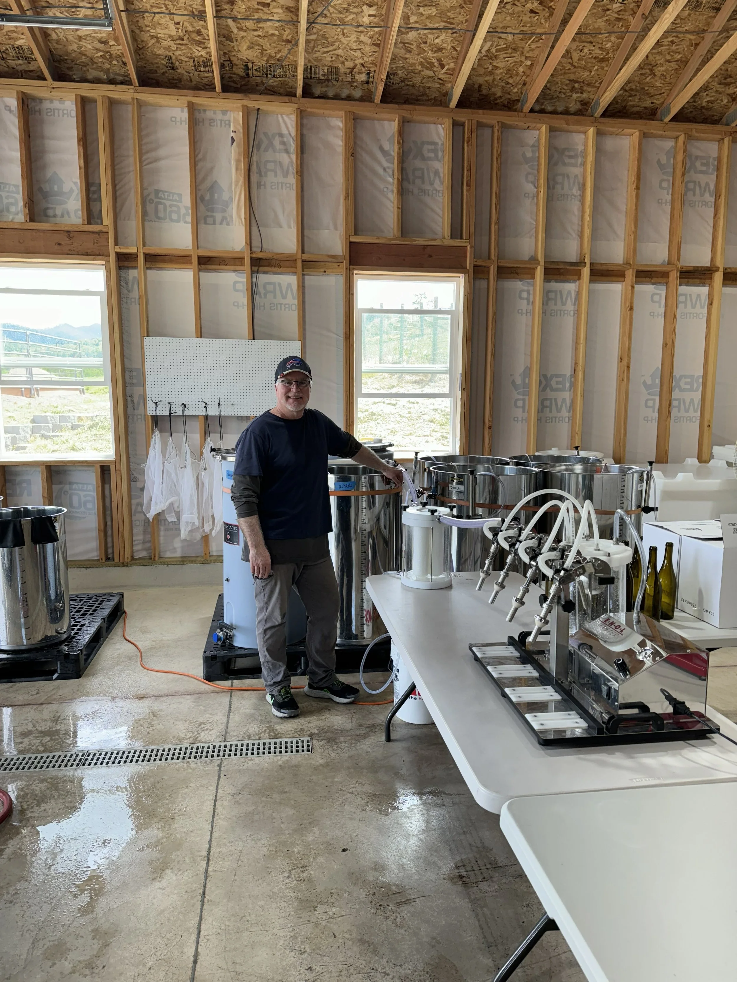 Man standing inside a building under construction, surrounded by brewing equipment, with water on the floor and wooden framing in the walls.