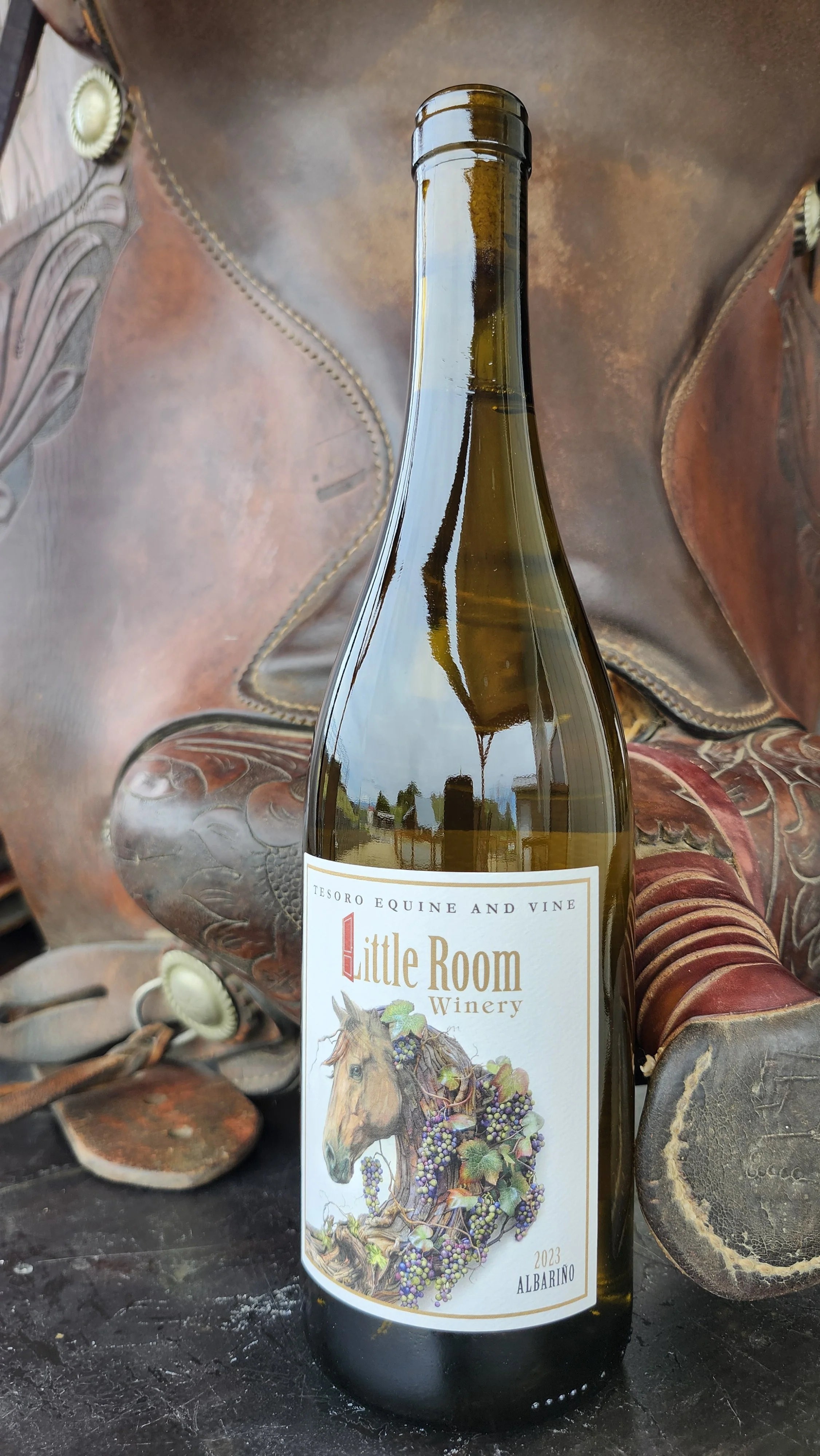 A wine bottle from Little Room Winery, labeled 2023 Albariño, placed next to a horse saddle with intricate stitching and embossing.