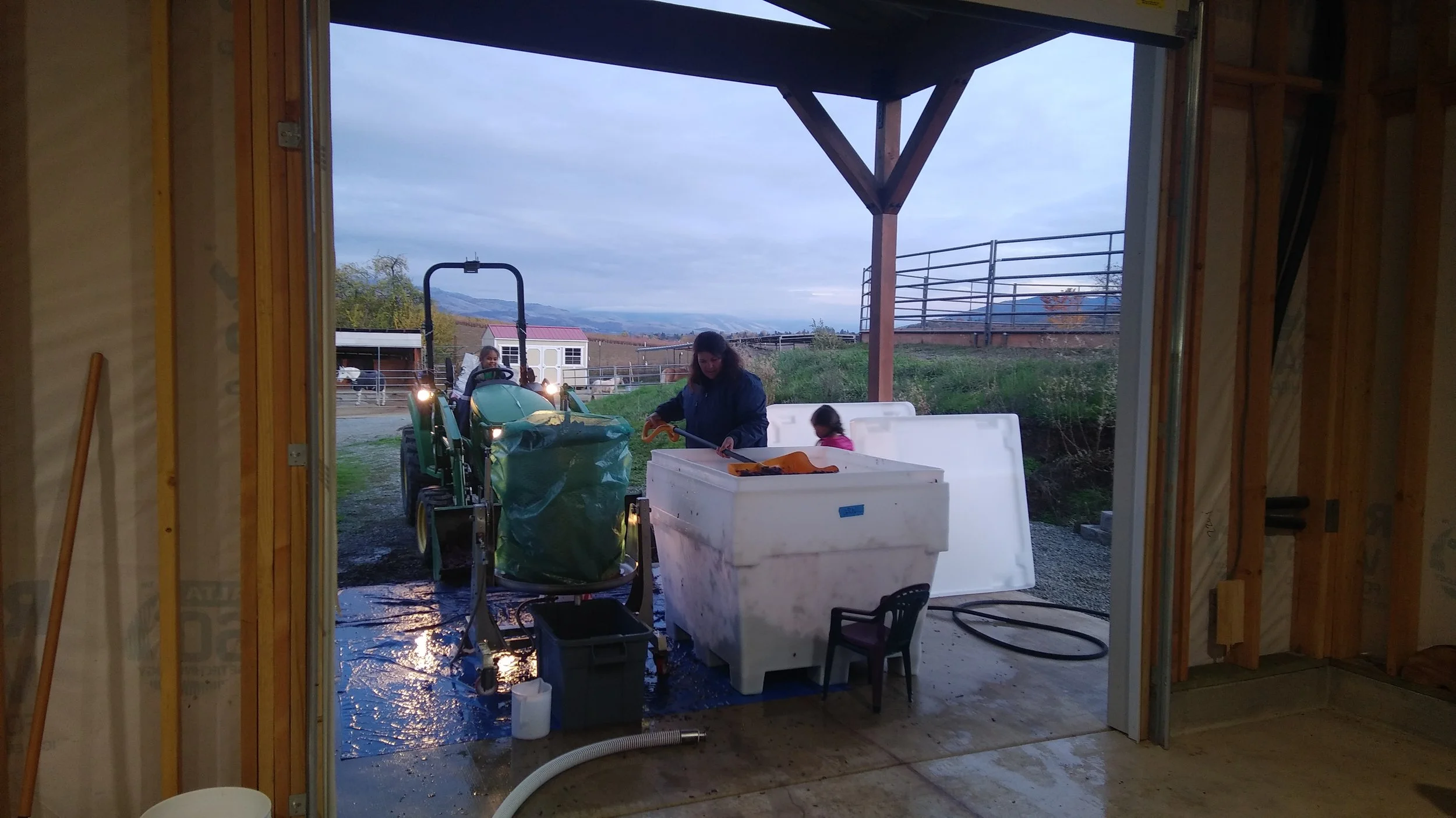 View from inside a winery building looking out onto the crush pad with a family. A woman and two children are in the yard, one girl on a green tractor and another girl helping her.  The sky is cloudy and distant mountains are visible.