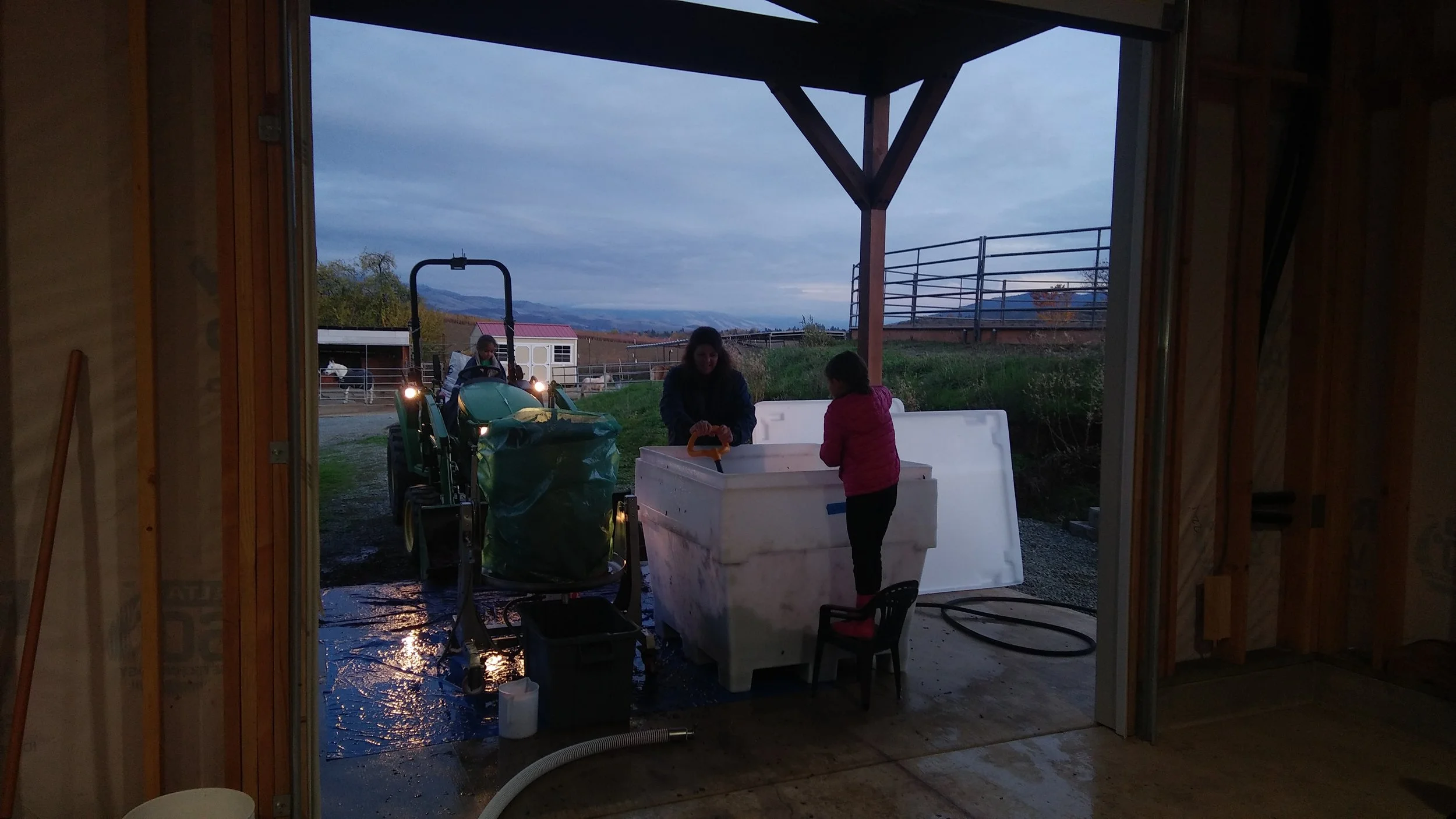 View from inside through the winery building opening showing two children on a farm, with a tractor outside, and a landscape of hills and cloudy sky in the background.