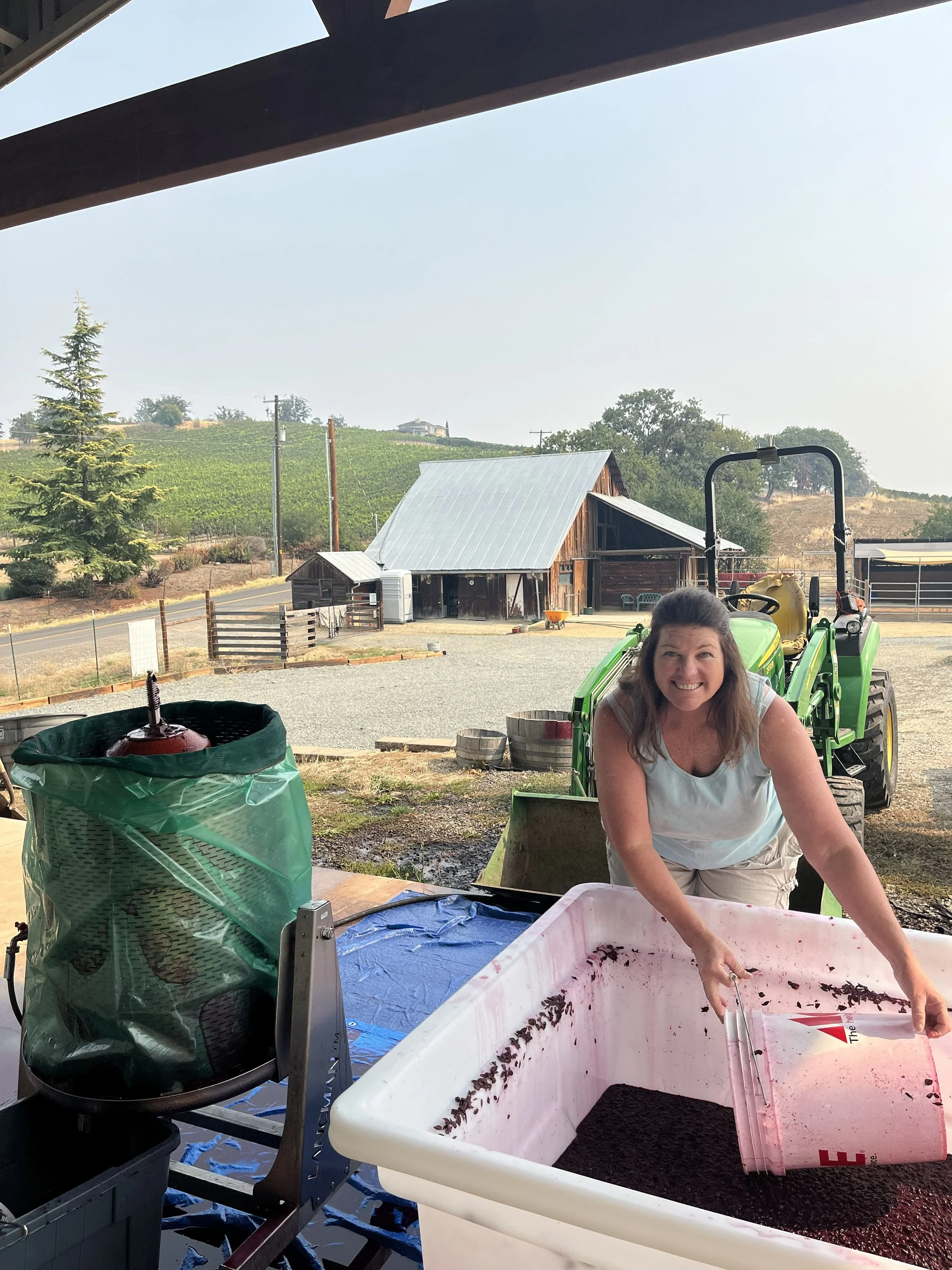 Woman smiling and working with a large container of crushed grapes outdoors, surrounded by farm buildings, vineyards, and hilly landscape.