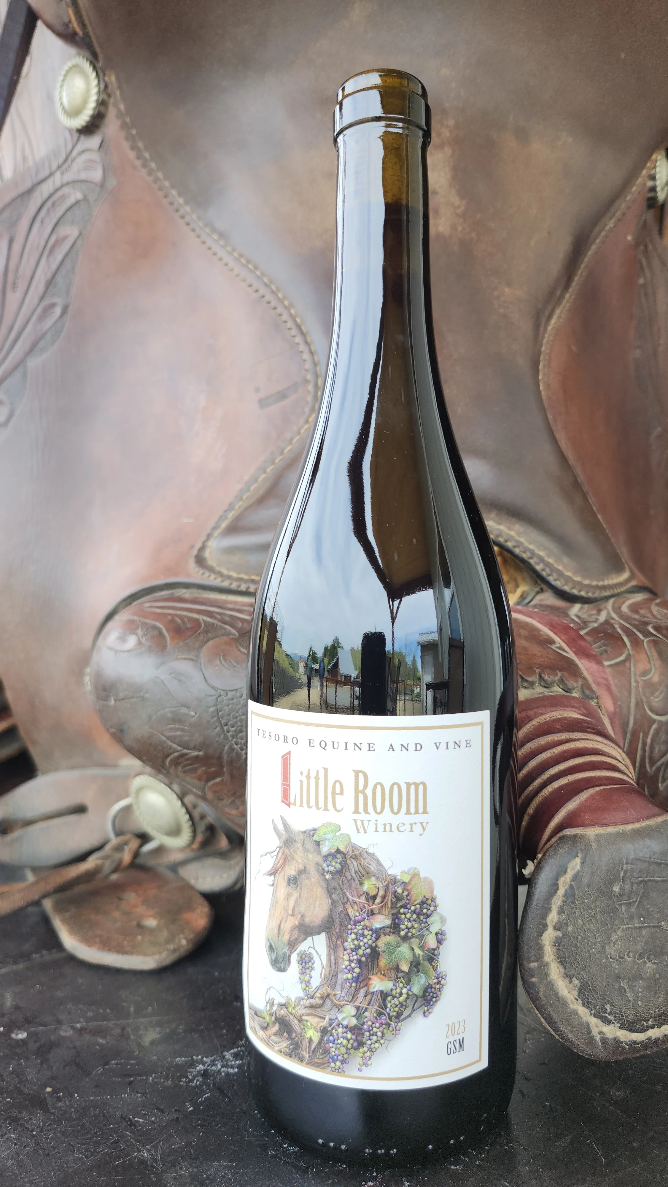 A wine bottle from Little Room Winery with a label featuring a horse surrounded by grapes and vine leaves, placed on a rustic wooden surface.