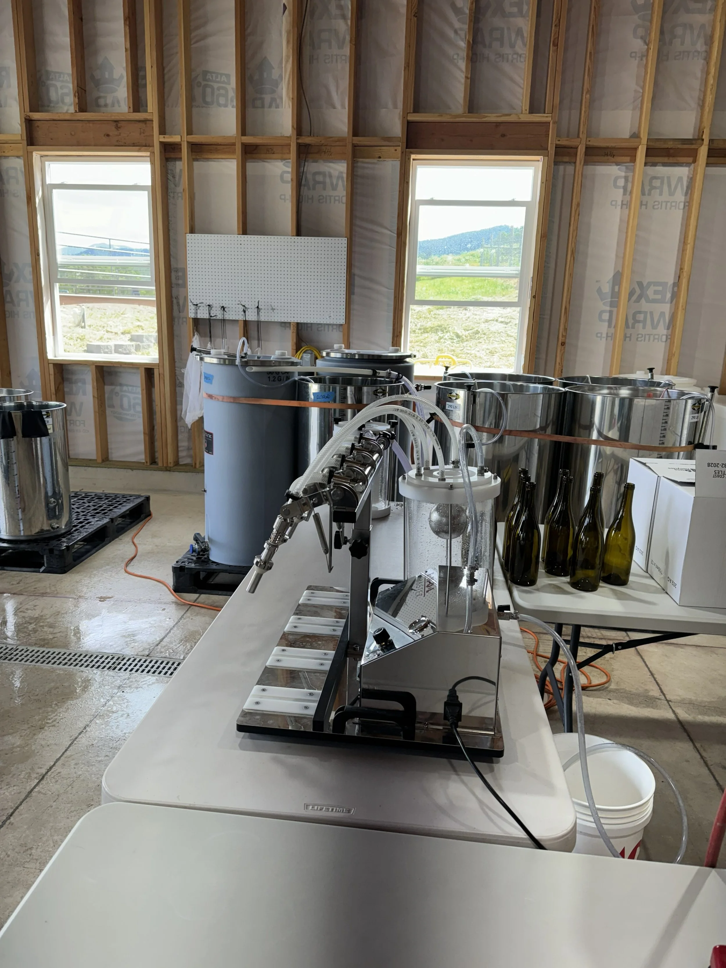 Interior of a building under construction with two windows, exposed wooden framing, and brewing equipment on a table.