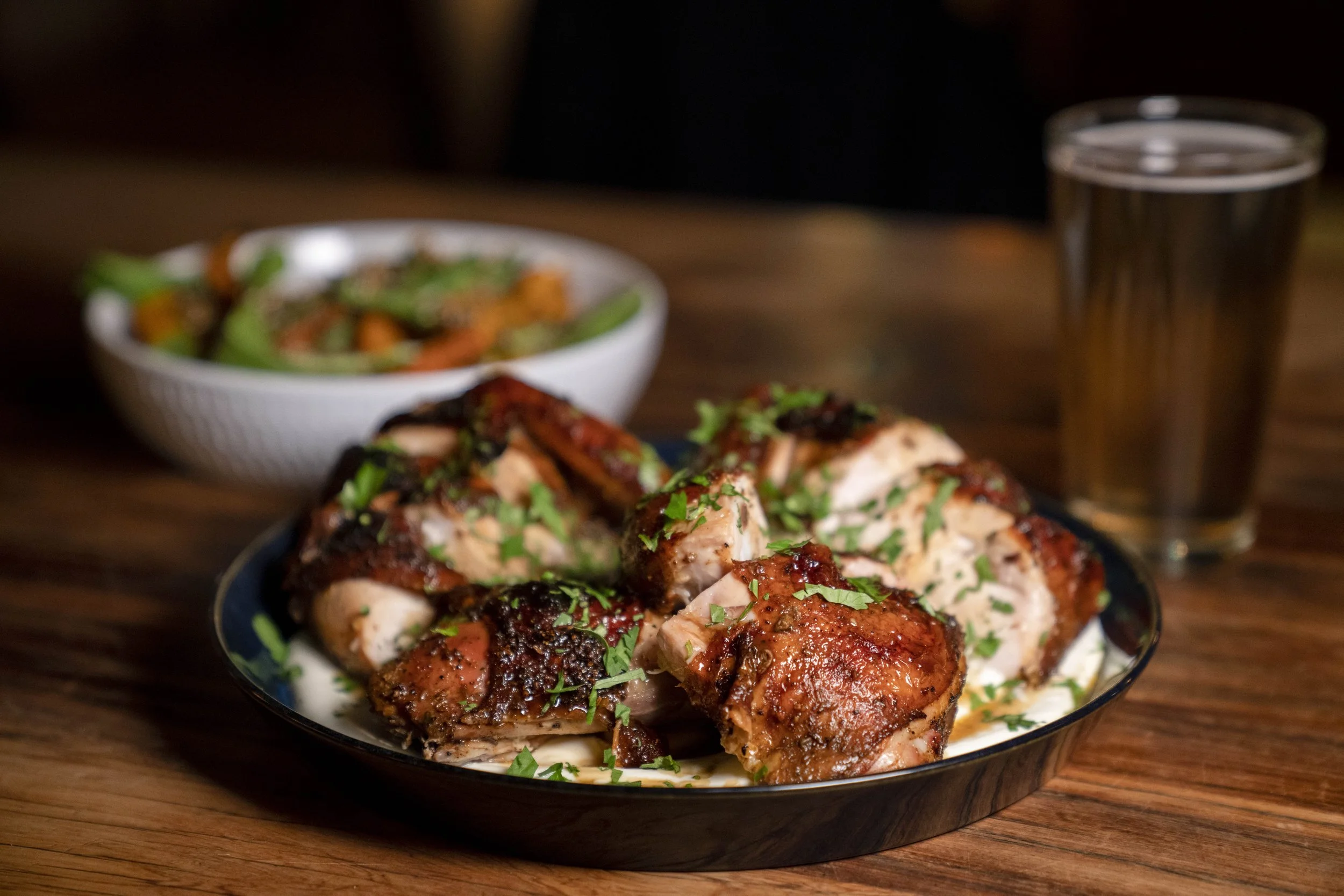Grilled chicken pieces garnished with chopped parsley served in a bowl, with a side salad and a glass of soda in the background.