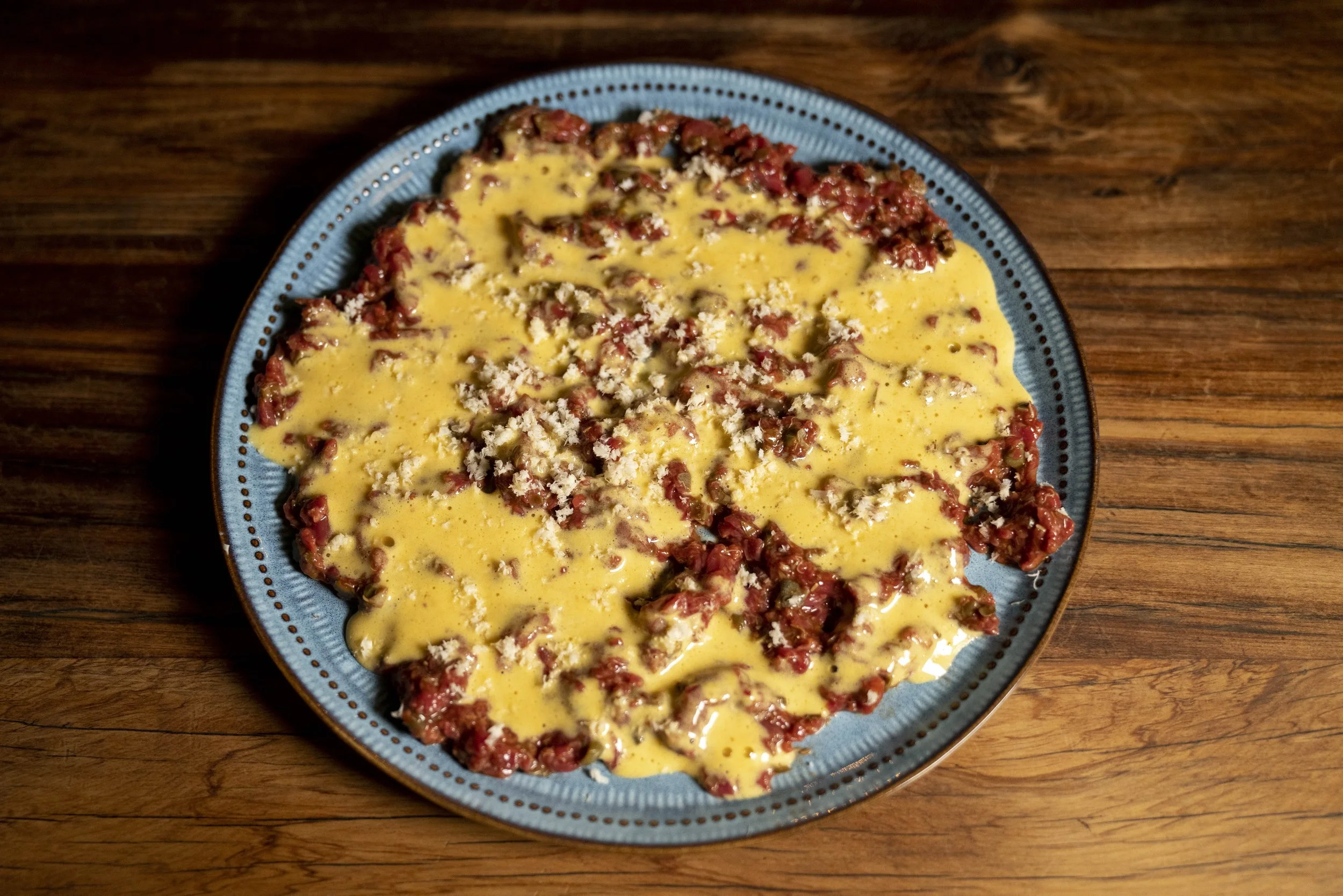 A blue plate with a meal of ground beef, cheese, and sauce on a wood table.