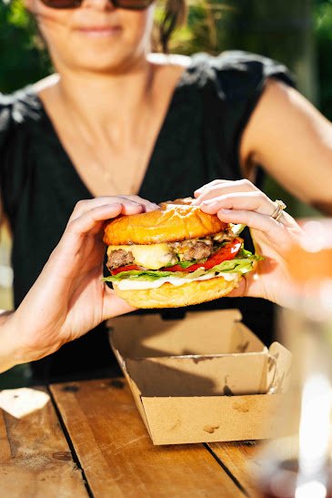 A woman holding a large cheeseburger with lettuce, tomato, and cheese, outdoors at a wooden table.