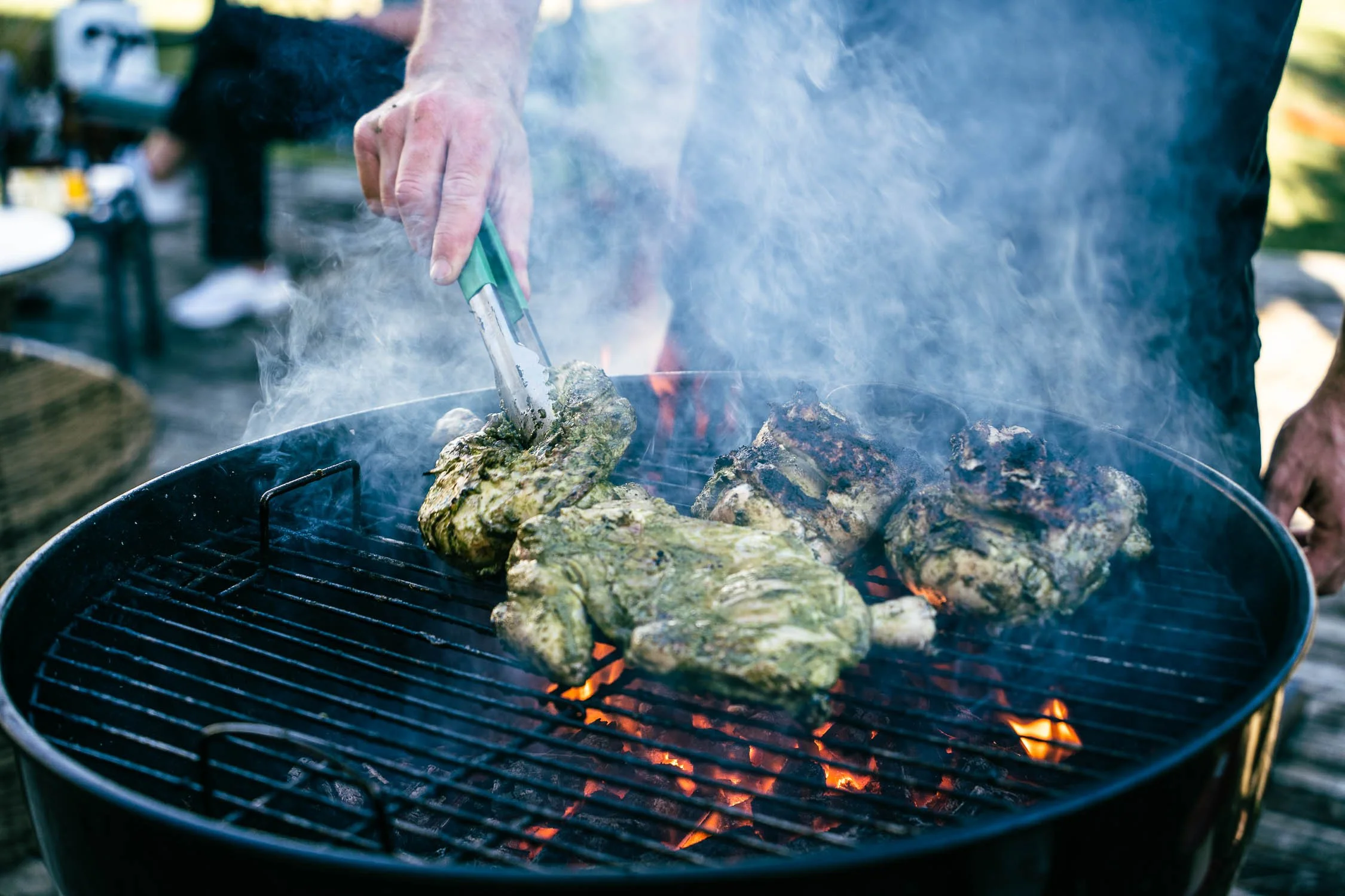 Person using tongs to handle a large piece of meat wrapped in foil on a charcoal grill with flaming coals and smoke.