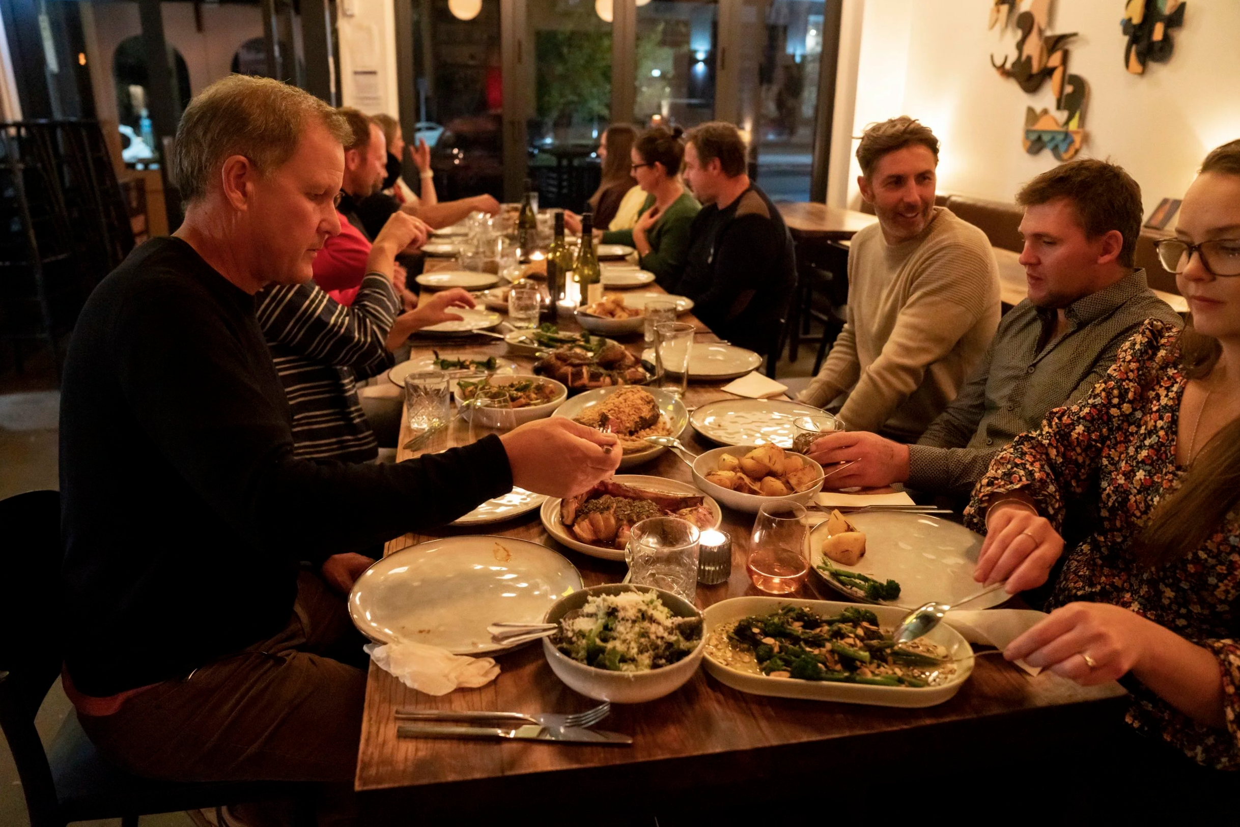 Group of people sitting at a long dinner table in a restaurant, enjoying a meal together with various dishes, including salads and cooked meats, in a warmly lit setting.