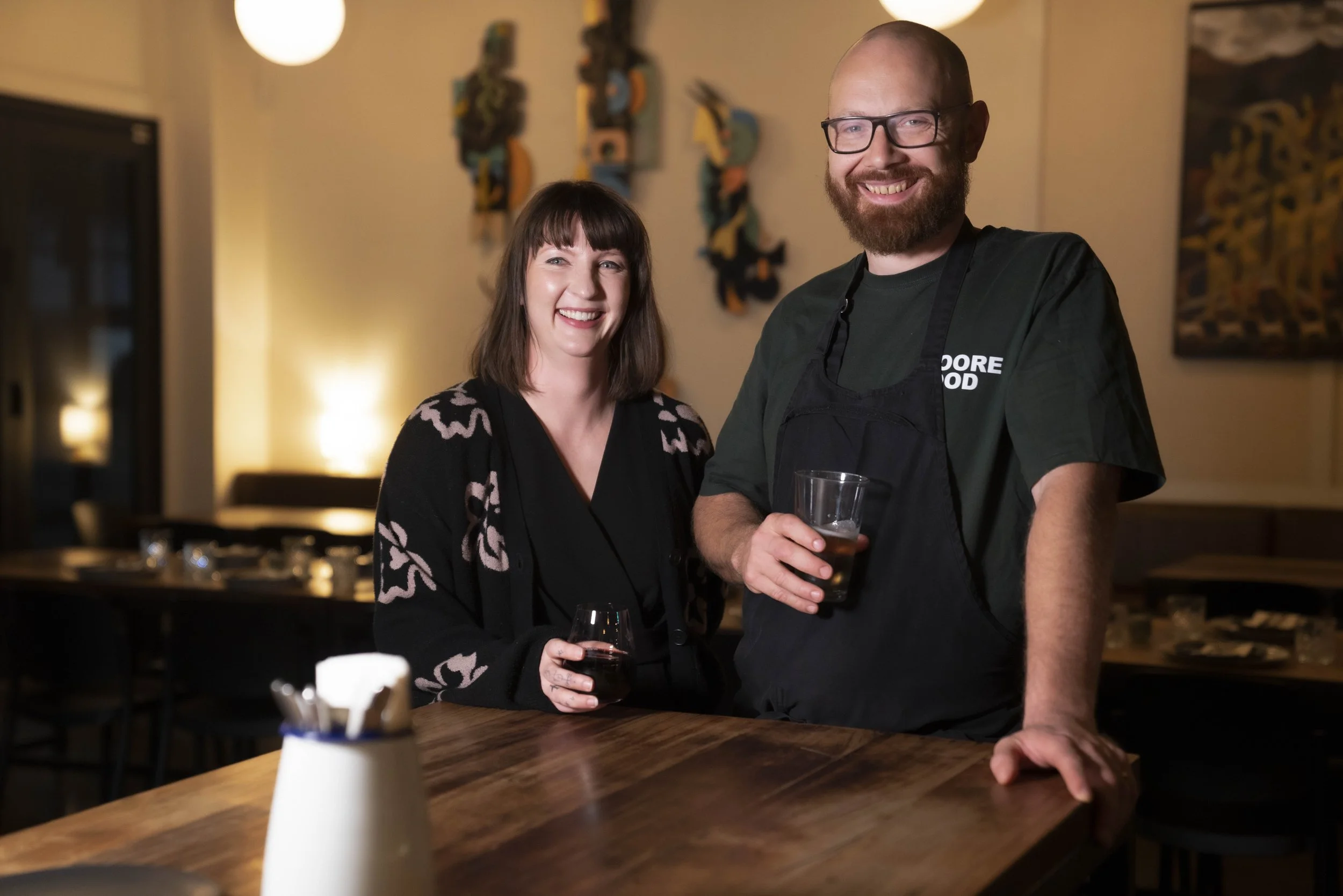 A smiling woman with brown hair and a man with glasses and a beard, both holding glasses of beer, standing in a dimly lit room that appears to be a restaurant or bar.