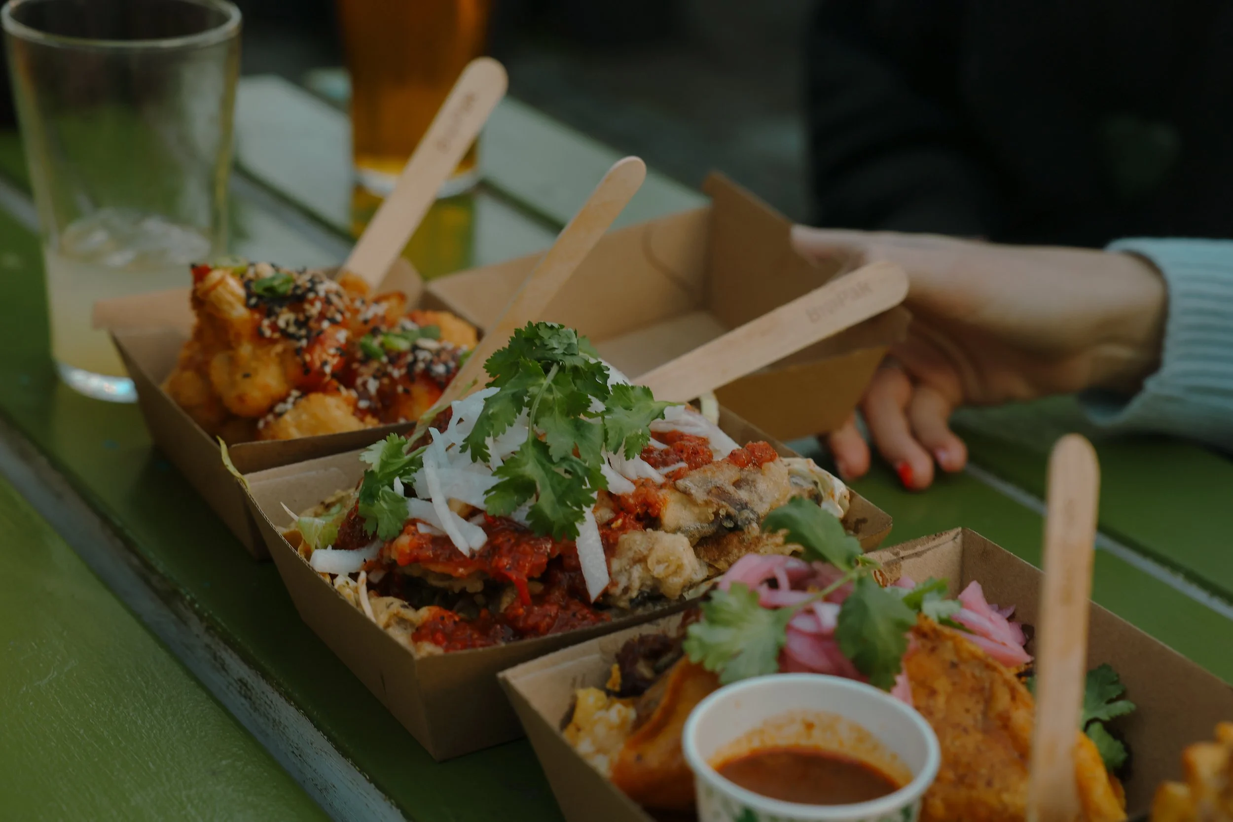 Assorted Asian dishes in brown takeout containers on a green table, with small cups of dipping sauces and beverages.