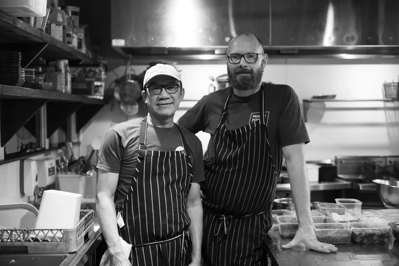 Two male chefs in aprons standing in a commercial kitchen, smiling at the camera.