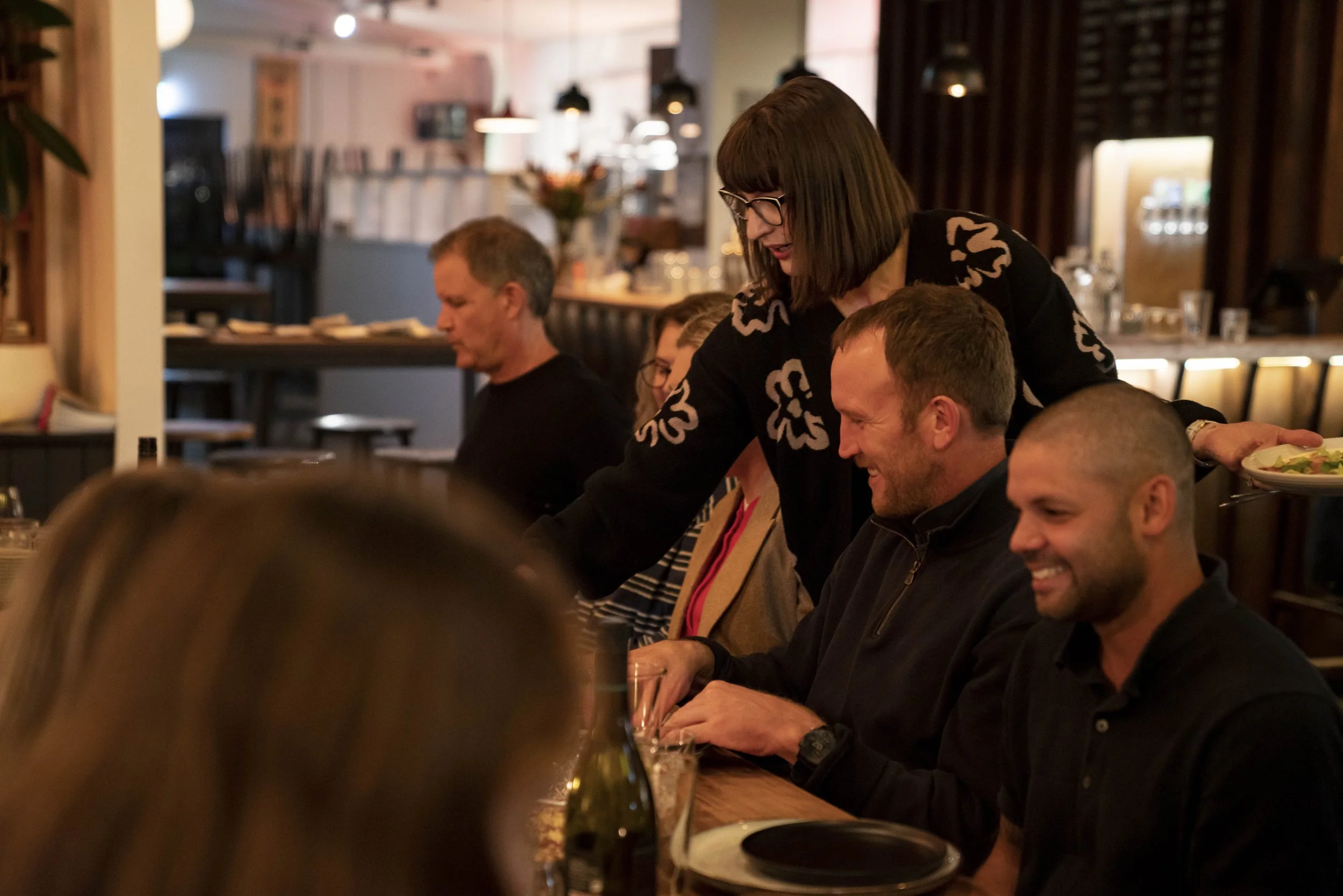 People sitting at a table in a cozy restaurant; a woman with glasses serving food to a man smiling near the front, with other diners visible in the background.