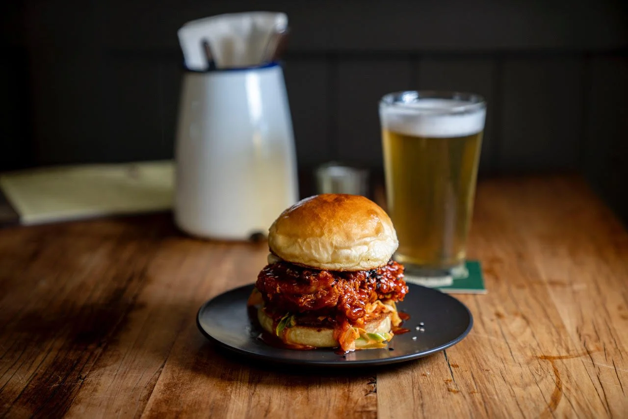 A plate with a fried chicken sandwich on a bun, served with a side of lettuce and sauce, accompanied by a glass of pale beer on a wooden table