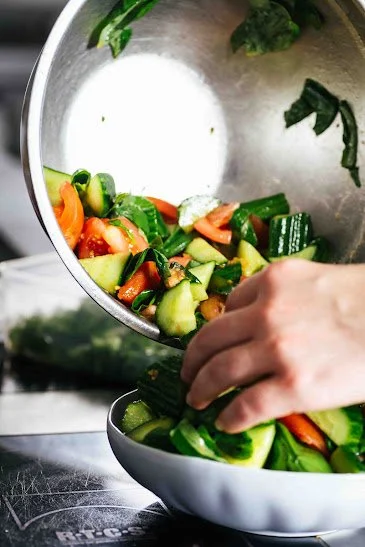 Hand pouring chopped cucumbers, carrots, and greens into a bowl in a kitchen.
