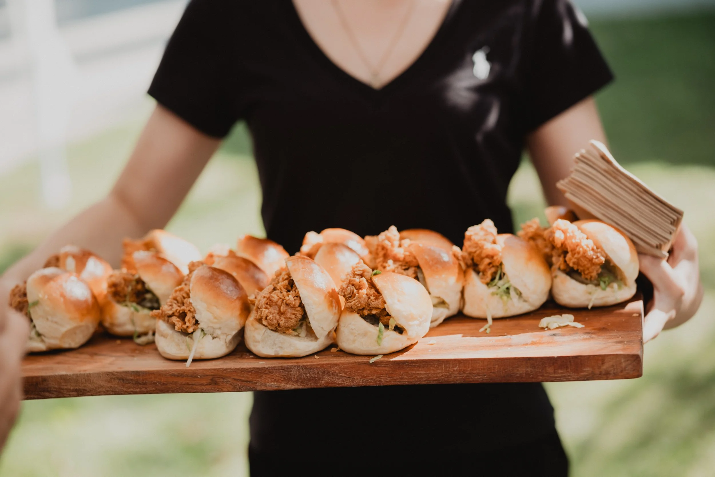 A person holding a wooden serving board with a row of small sausage sandwiches with buns, fried meat, and shredded lettuce inside, outdoors in a green setting.