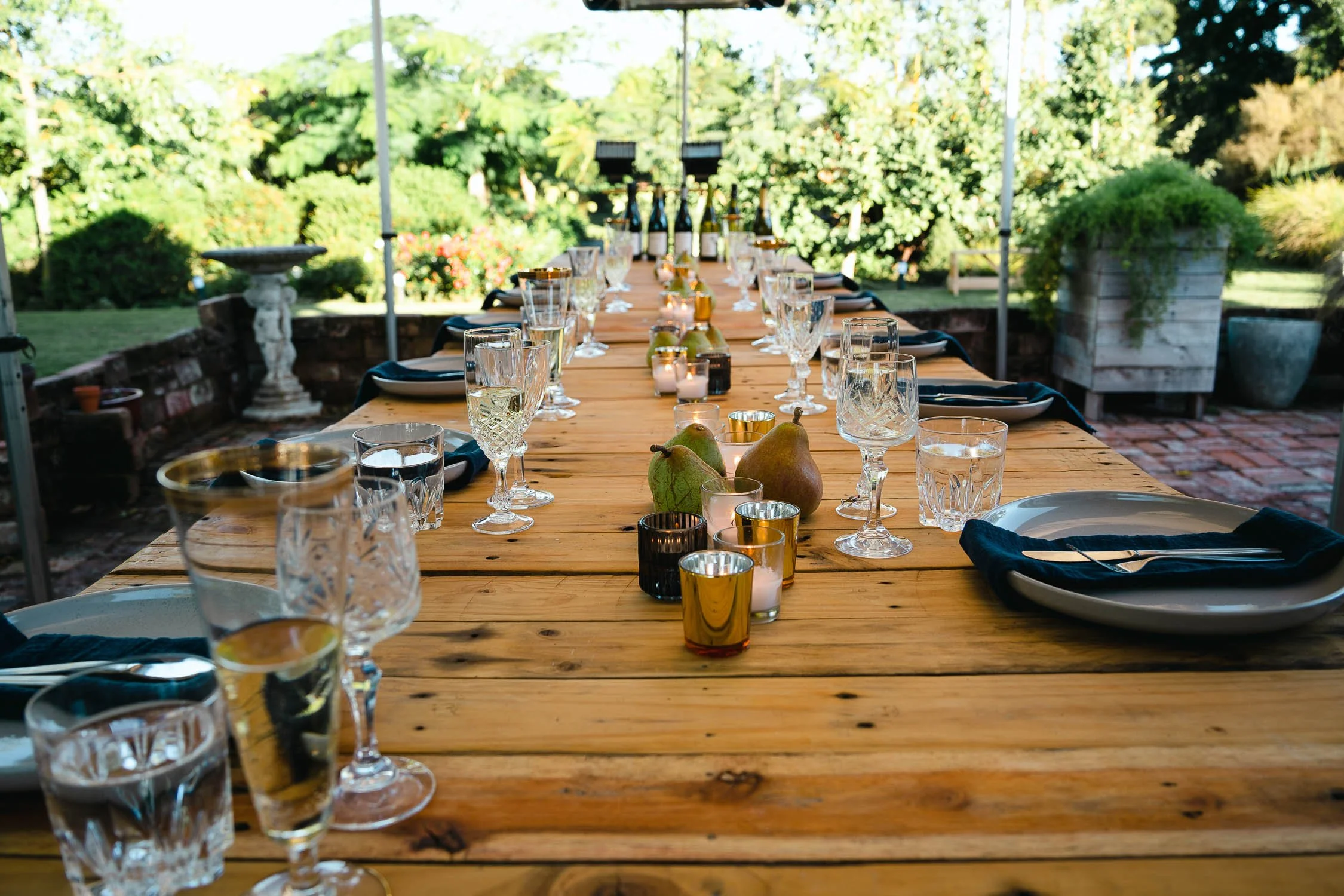 Long wooden outdoor dining table set with glassware, candles, pears, and black napkins, surrounded by lush green garden with trees and plants.
