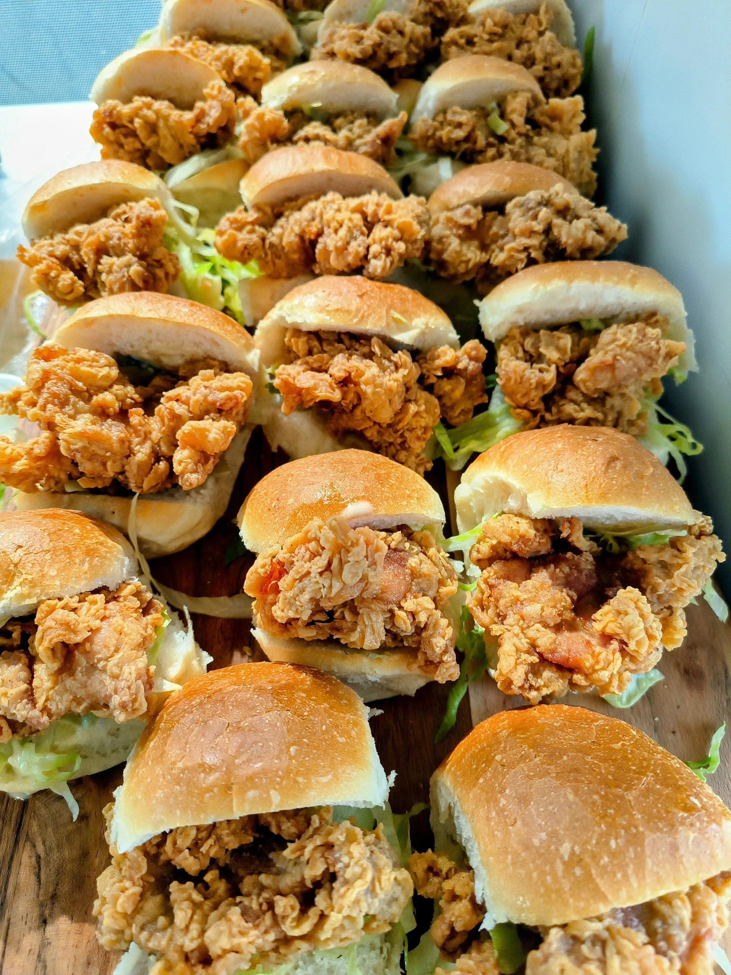 Close-up of various fried chicken sliders on a wooden platter, with some containing lettuce and sauce.