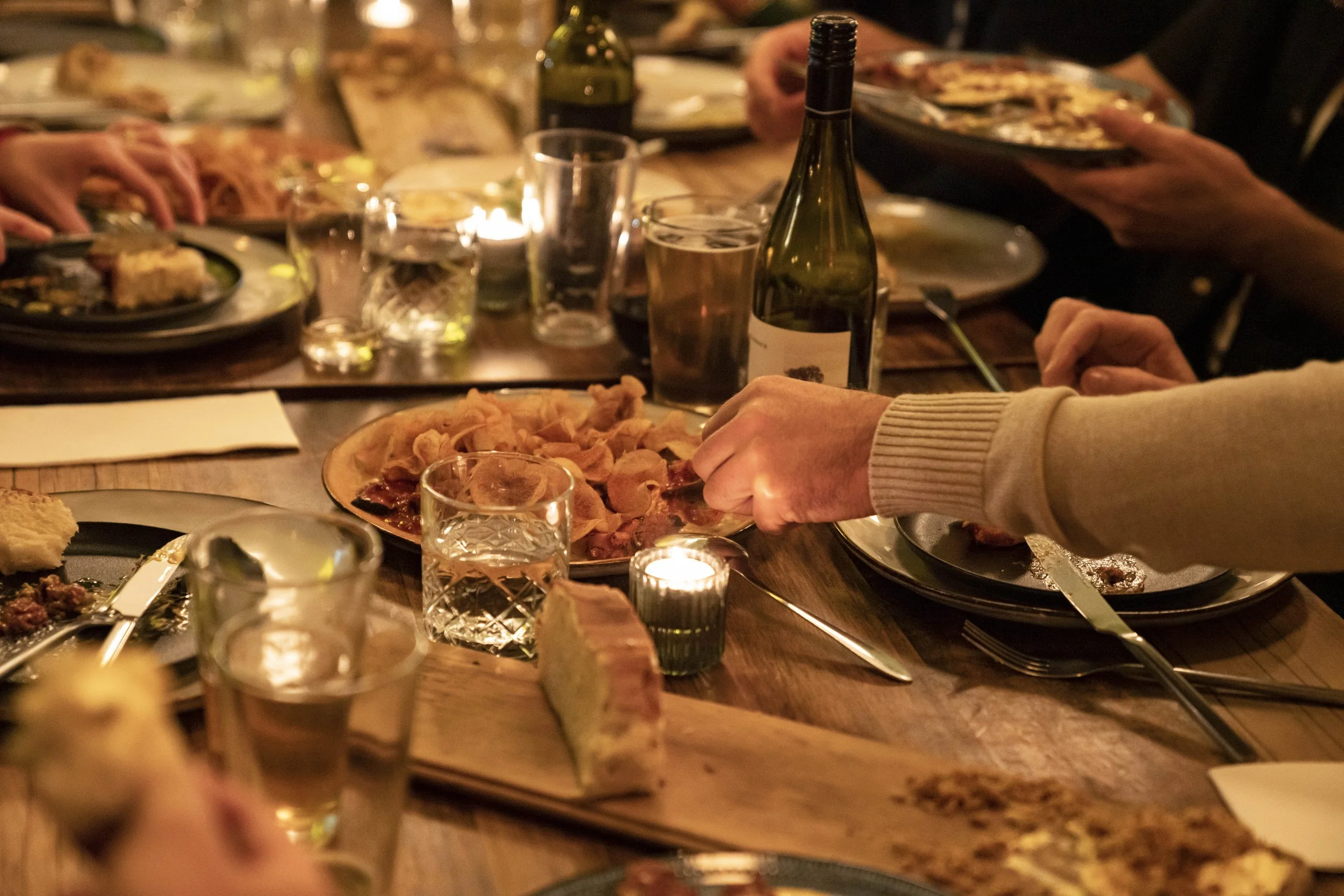 A dinner gathering in a dimly lit restaurant with numerous plates of food, glasses, bottles of wine, and candles on a wooden table. People are reaching for food and serving themselves.