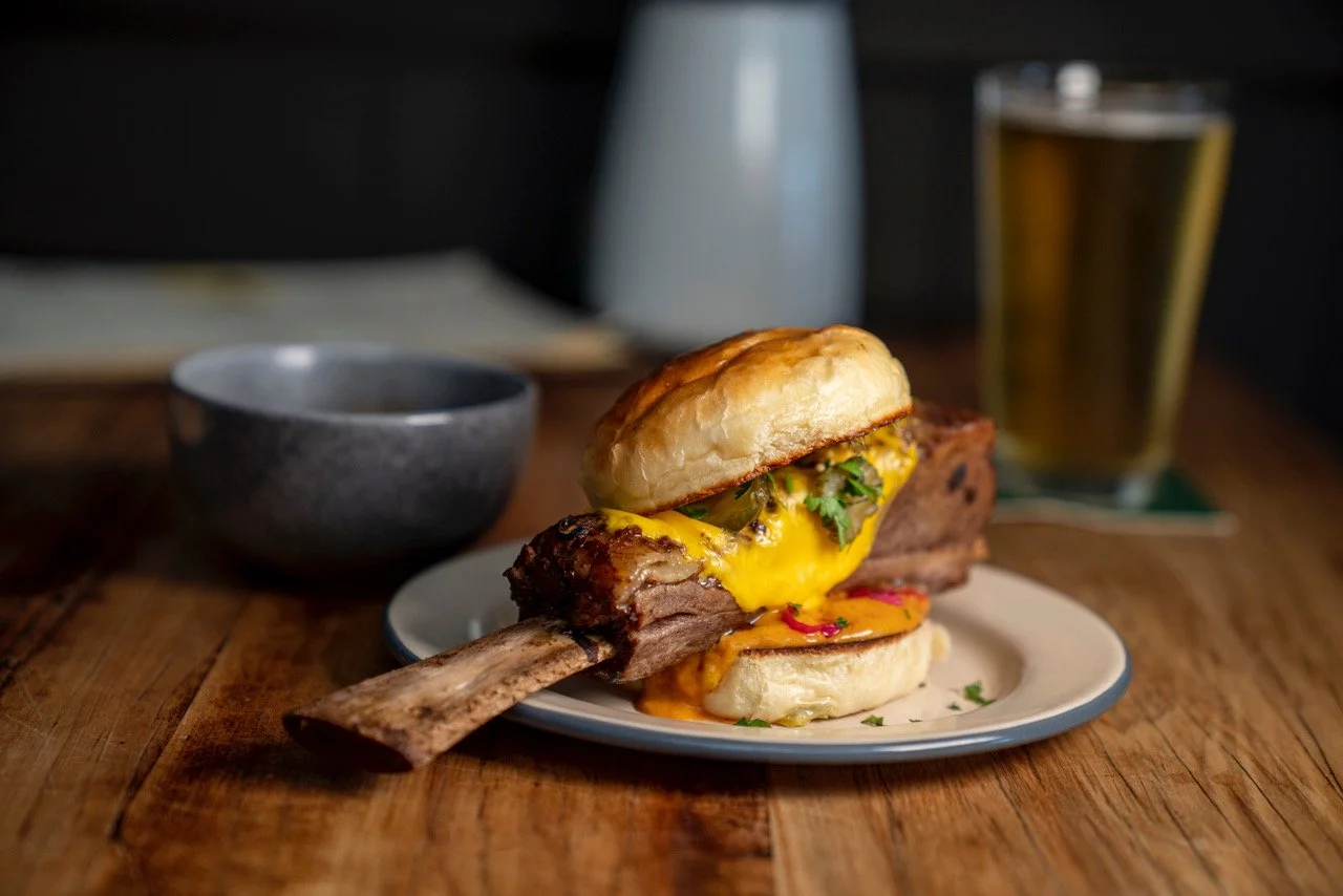 A cheeseburger with a beef patty, melted cheese, and a bun placed on top, served with a side of sauce and garnished with herbs on a white plate. In the background, there is a glass of beer, a small black bowl, and a white pitcher on a wooden table.