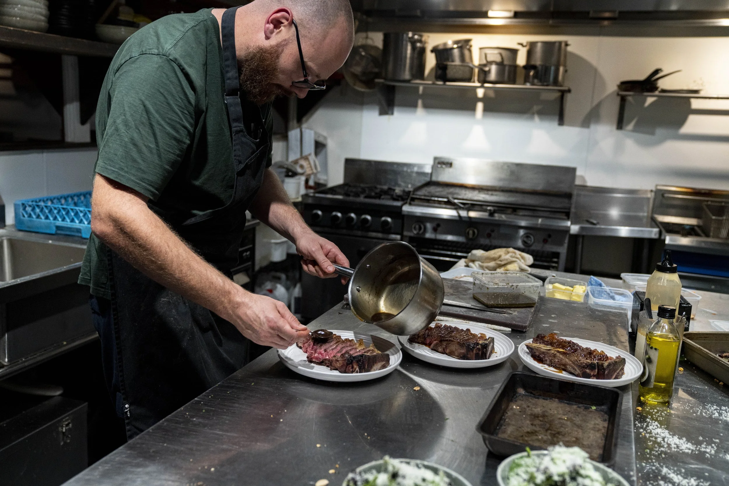 A man with a beard is plating cooked meats in a commercial kitchen. The kitchen has stainless steel appliances and shelves with cookware. The food is on paper plates on a stainless steel counter.