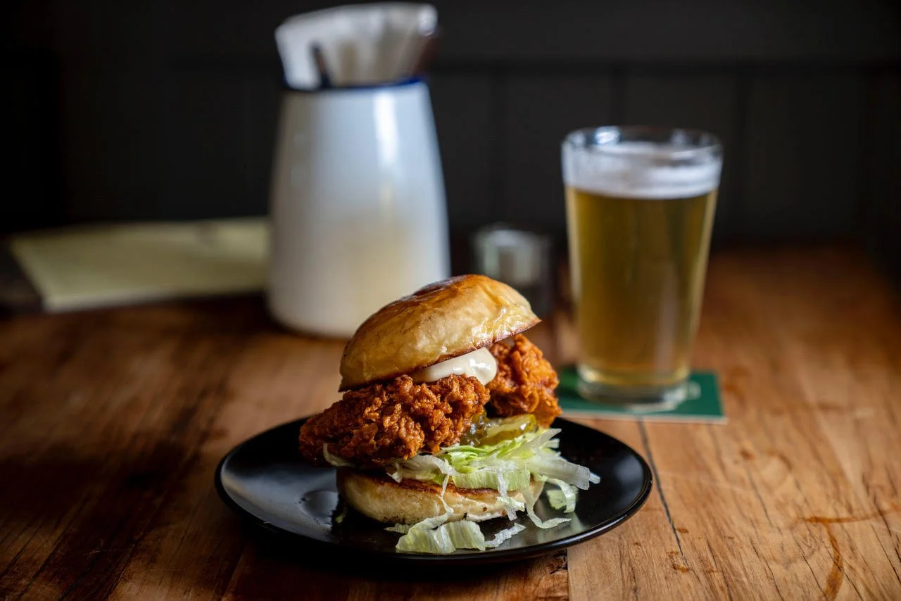 Fried chicken sandwich with lettuce and mayonnaise on a bun, served on a black plate. A glass of beer and a white container with utensils are in the background on a wooden table.