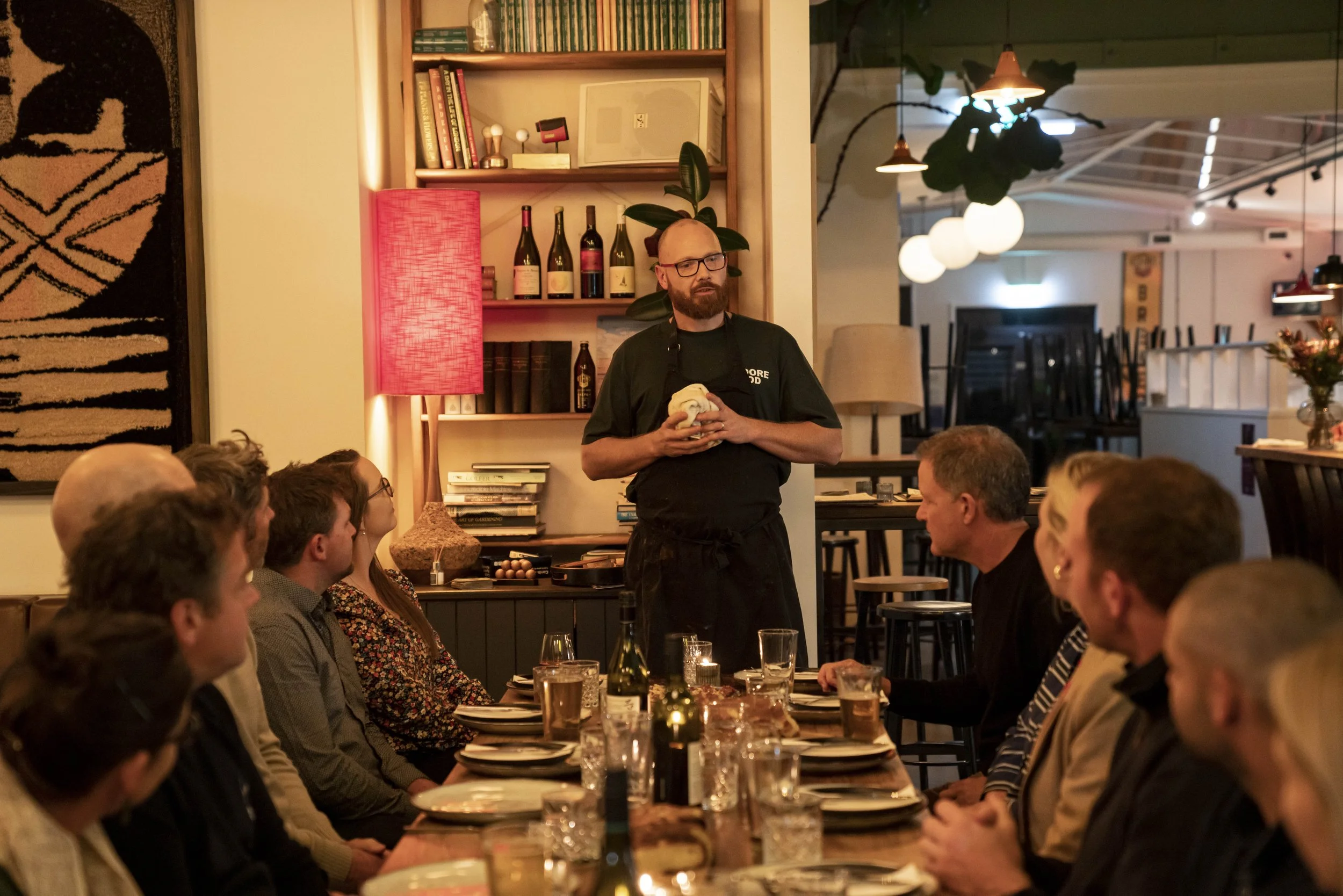A man with glasses and a beard, wearing a black shirt and apron, is standing in front of a group of people at a dining table, holding a cloth. The setting is a warmly lit restaurant or cafe with shelves of books and bottles of wine in the background,