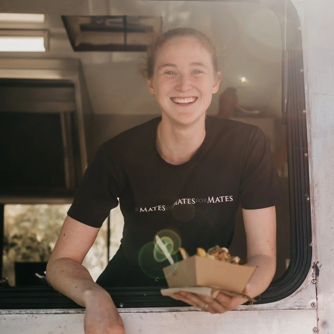 A smiling woman with brown hair tied back, wearing a black T-shirt that says 'MATES FOR MATES,' offers a food tray through a window.