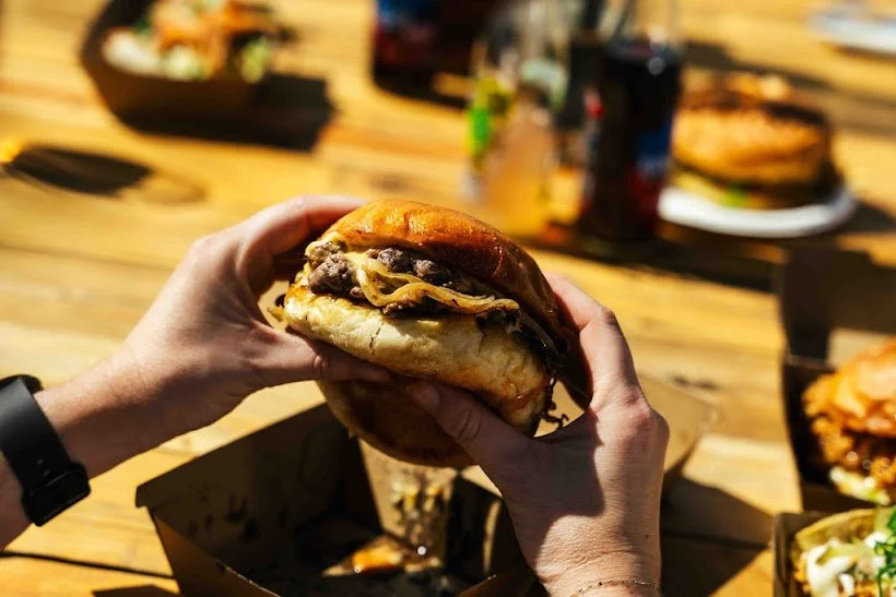 Person holding a cheeseburger with beef patty, cheese, and bun in a restaurant setting with other burgers and fries visible on a wooden table.