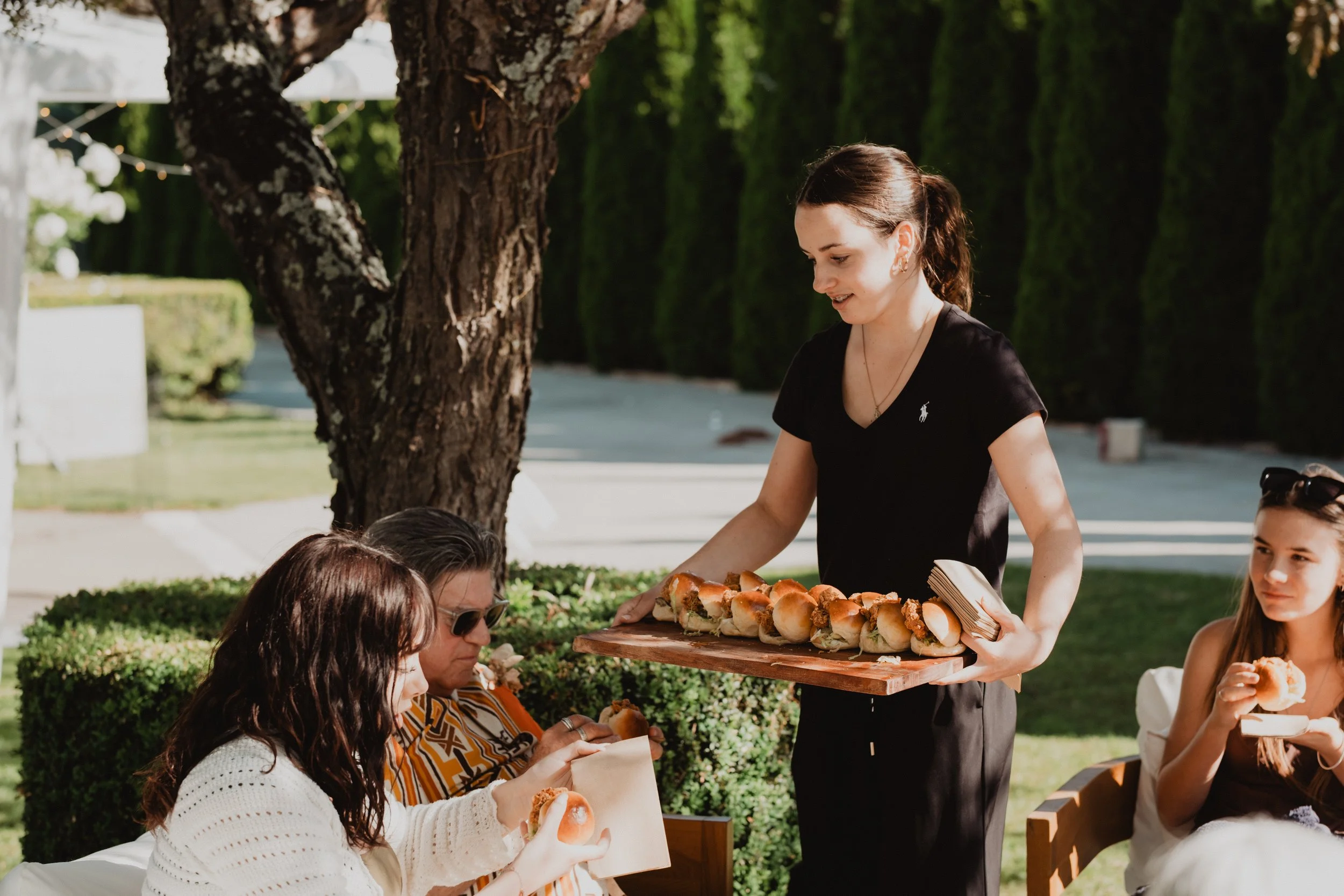 A woman in black shirt serving hot dog sliders to a group of people at an outdoor gathering under a tree.