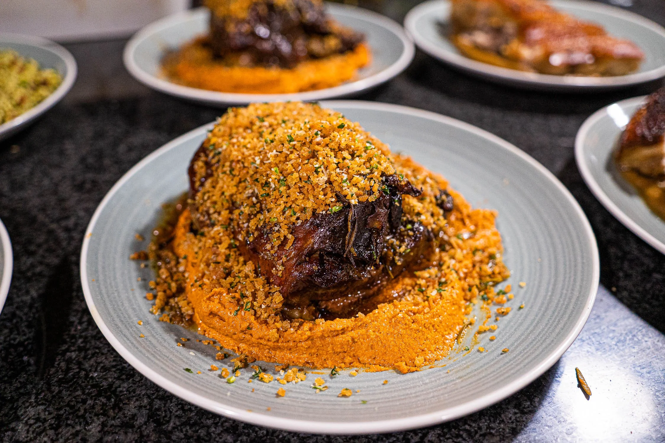 A plate with a mound of baked meat covered with tomato sauce and topped with crunchy seasoned breadcrumbs, with other plates of similar dishes in the background on a dark countertop.