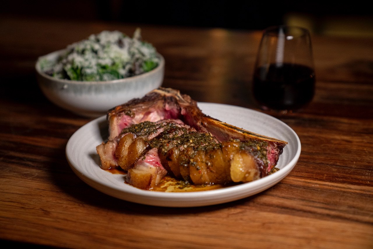 A plate of cooked lamb chop with sauce, a bowl of salad, and a glass of red wine on a wooden table.