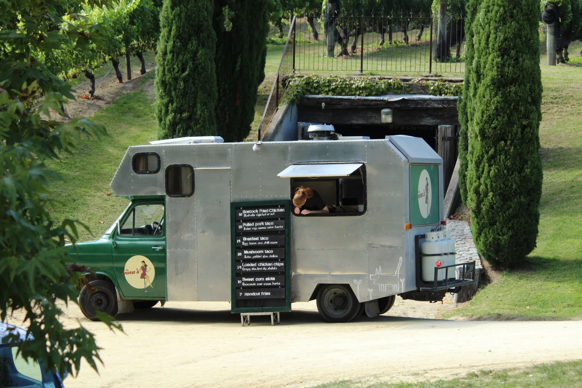 A food truck with a menu on the side, parked on a dirt surface, with two tall cypress trees and a grassy area in the background. There are animals grazing behind a black fence.