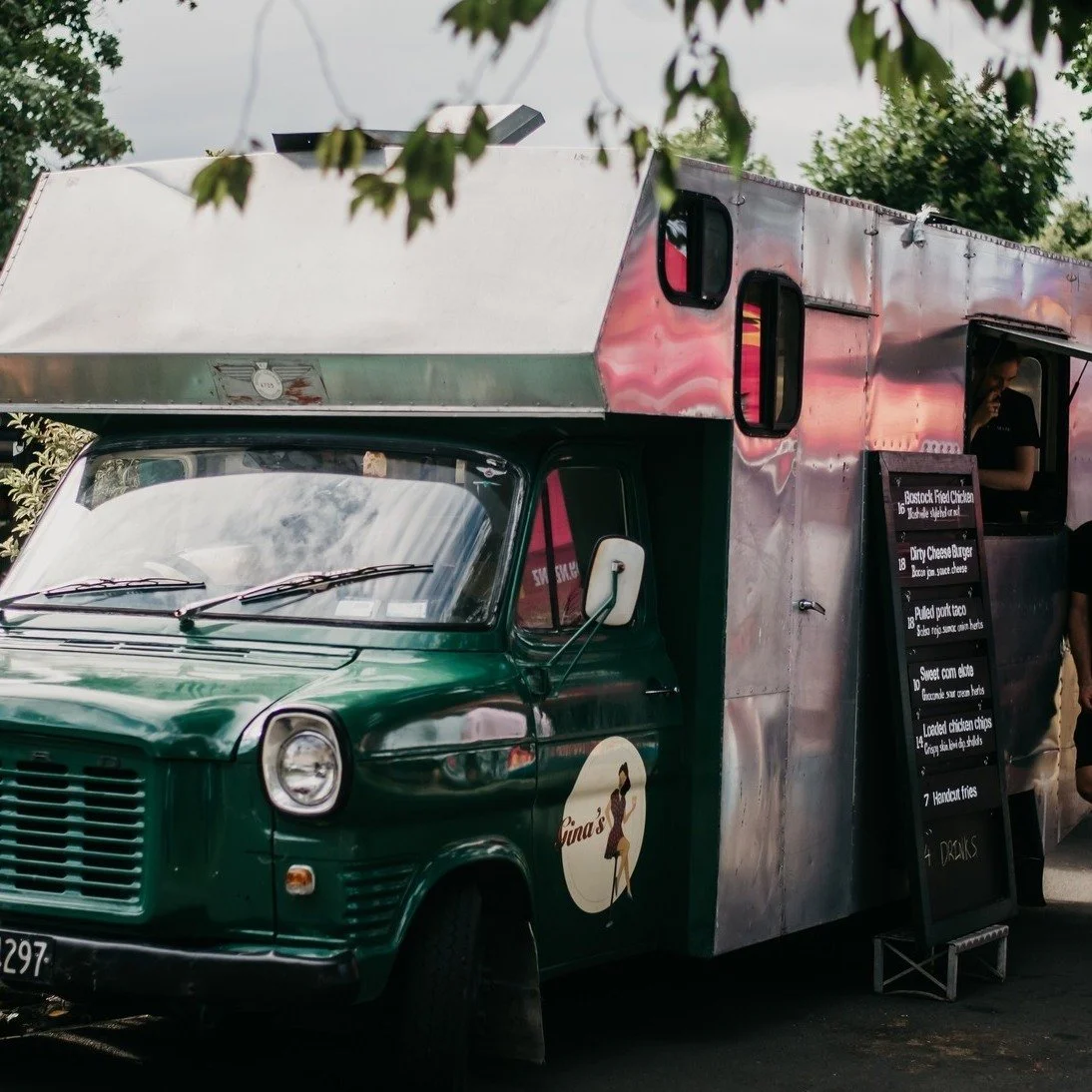 A green food truck with a metal upper section and a black menu board displaying various food items next to a small window where a person is standing.