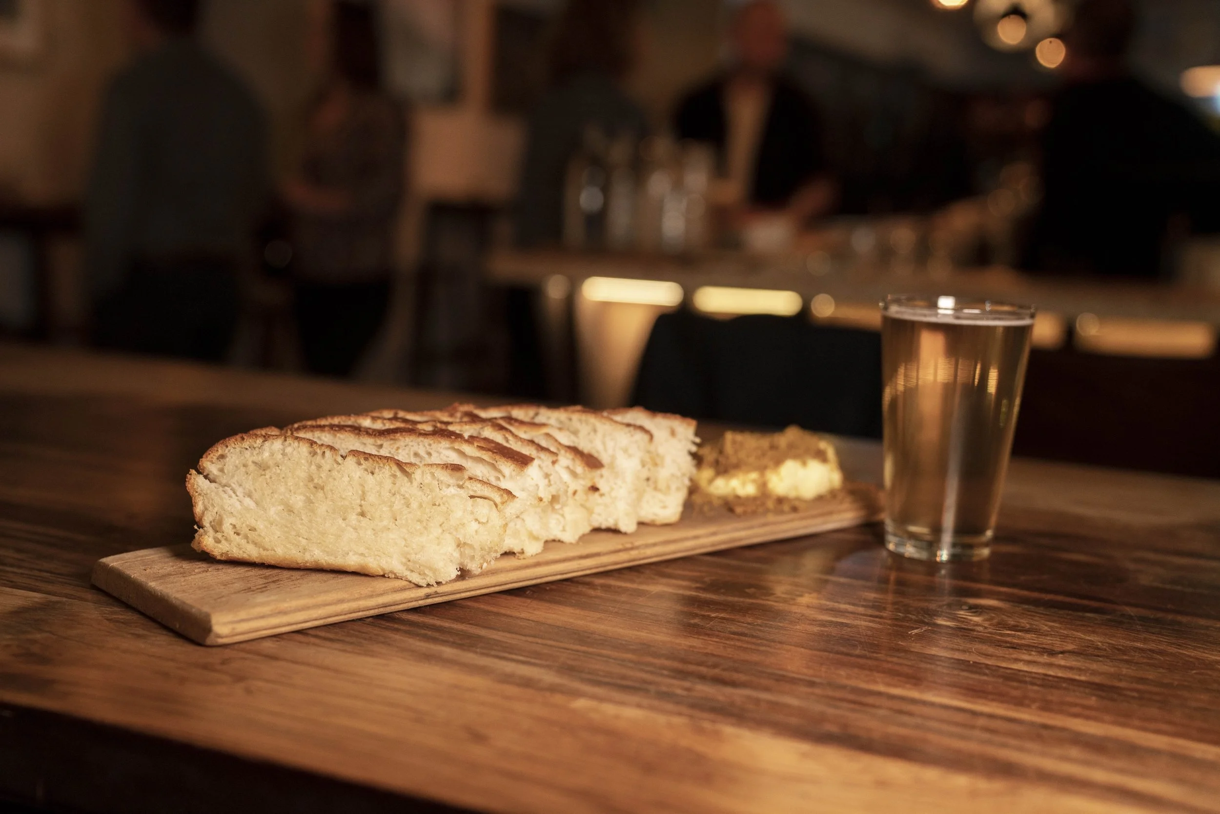 Sliced loaf of bread on a wooden cutting board with a glass of water or clear beverage beside it, in a dimly lit cafe or restaurant setting.