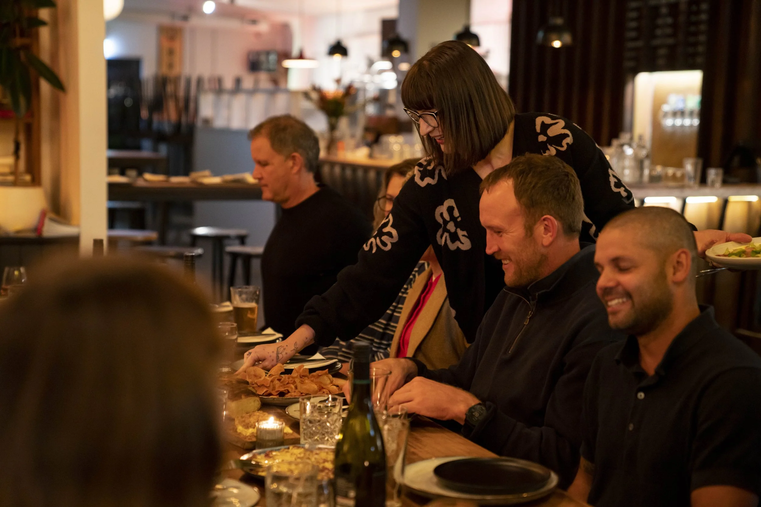 A group of people dining at a restaurant, with a woman serving food to others, seated at a long table with various dishes and drinks, warm lighting, and a cozy ambiance.